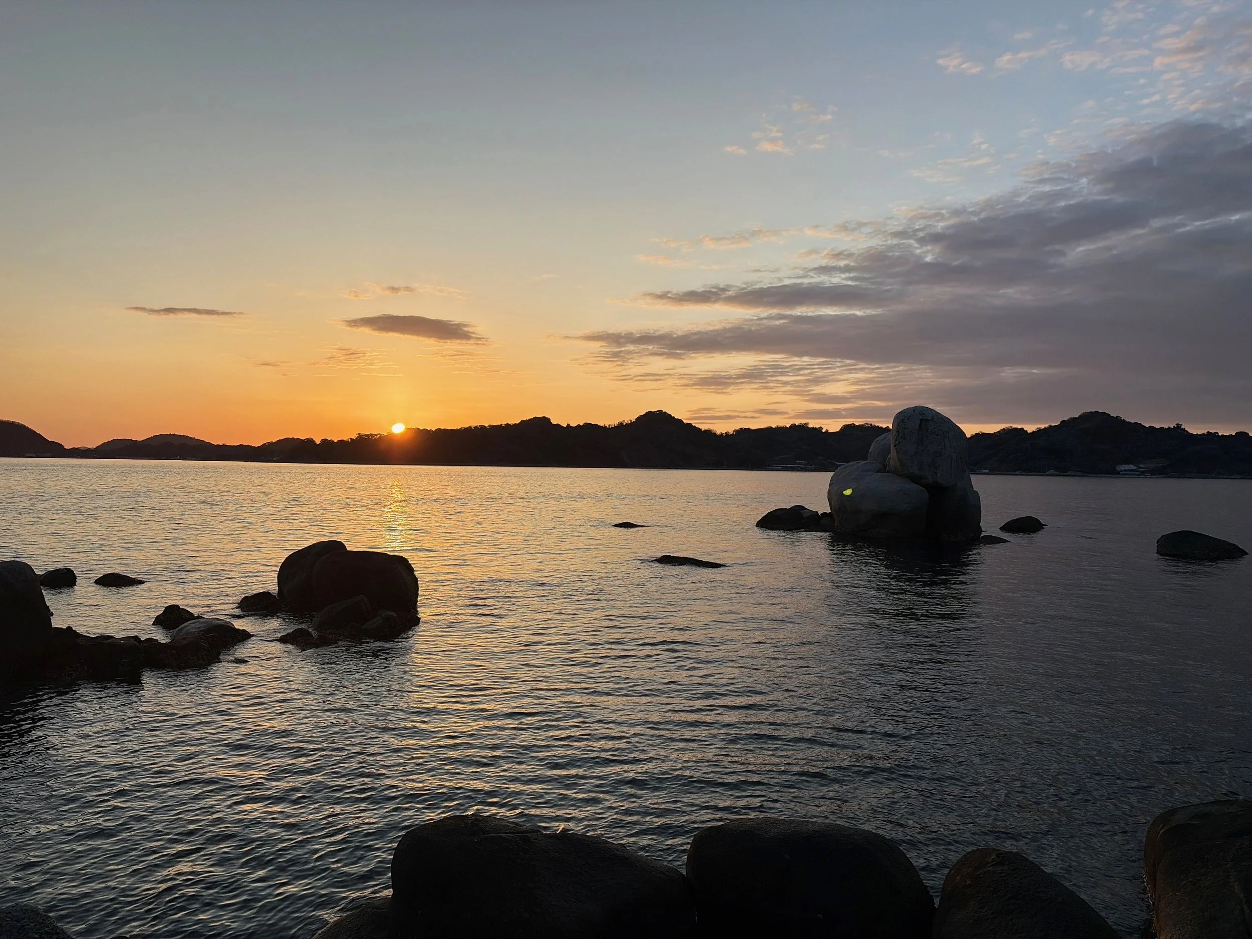 夕暮れの海を正面から望む風景です。水平線の近くに沈みかけた太陽があり、海面には黄金色の光が細長く反射しています。遠くには低い山々や島々が黒いシルエットとなって連なり、右側の海には大きな丸い岩がいくつも並んでいます。手前にも大きな岩があり、穏やかな波がその周りを静かに揺らしています。空は橙色から青灰色へ変わり、雲が広がる幻想的な夕景です。