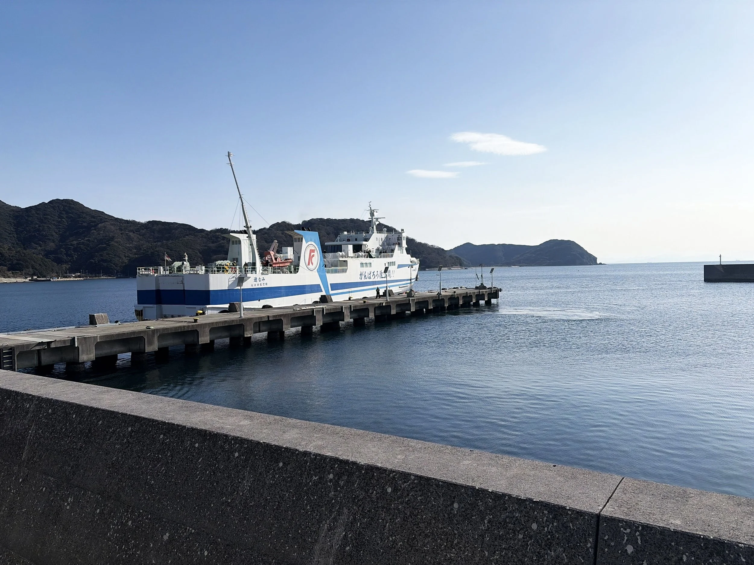 青い海に面した港に、一隻の白と青のフェリーが停泊しています。船は長い桟橋につながれ、船体には大きく文字が描かれています。手前には黒いコンクリートの防波堤があり、その向こうに穏やかな海が広がっています。海の奥には緑に覆われた山々が連なり、右側には小さな防波堤も見えます。青空の下、静かな港と船が印象的な風景です。