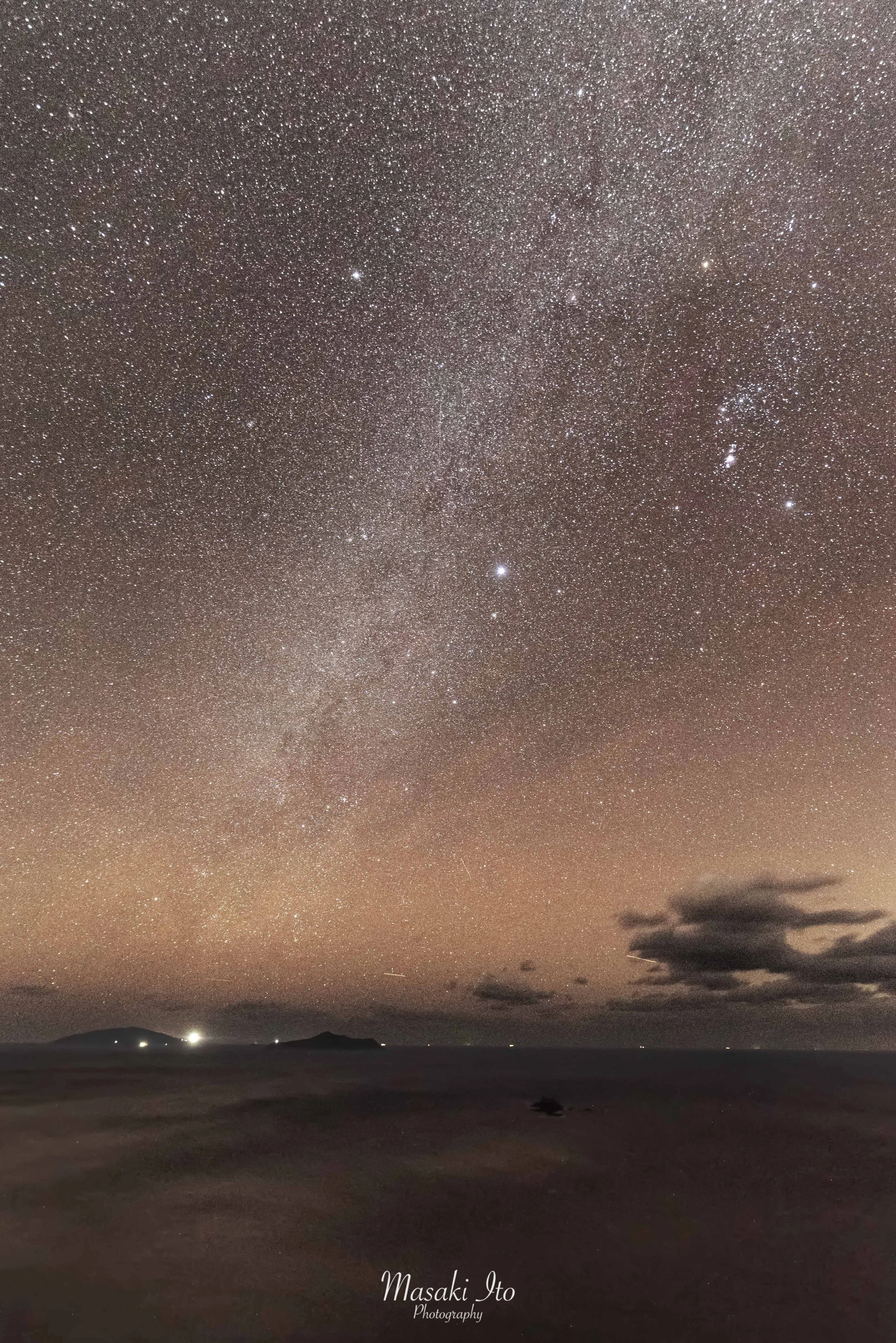 夜の海辺から見上げた満天の星空です。空一面に無数の星が散りばめられ、中央付近には淡く白い天の川が縦に伸びています。右側にはひときわ明るい星々が集まり、星座のような形を作っています。水平線には小さな島や山の影が浮かび、いくつかの明かりが静かに輝いています。海と夜空の境界が溶け合う、幻想的で静かな夜の風景です。