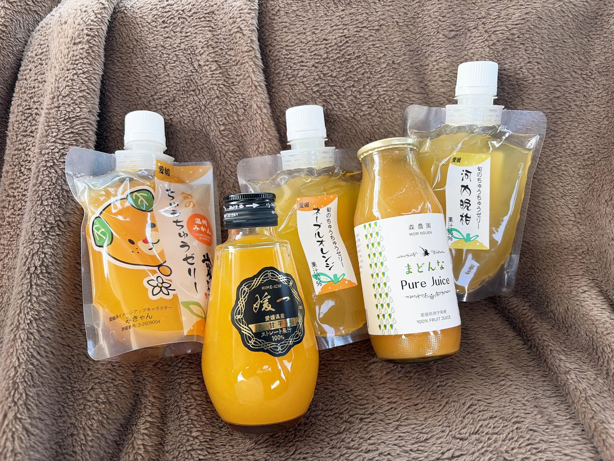 茶色の柔らかな毛布の上に、愛媛県産の柑橘ジュースが5本並べられています。オレンジ色の液体が入ったパウチや瓶が中心で、前には「甘平」の100％ストレート果汁、右には「まどんな」のピュアジュースがあります。中央奥にも柑橘ジュースのパウチがあり、全体的に鮮やかなオレンジ色が目を引きます。愛媛らしい柑橘の豊かな味わいを感じさせる写真です。