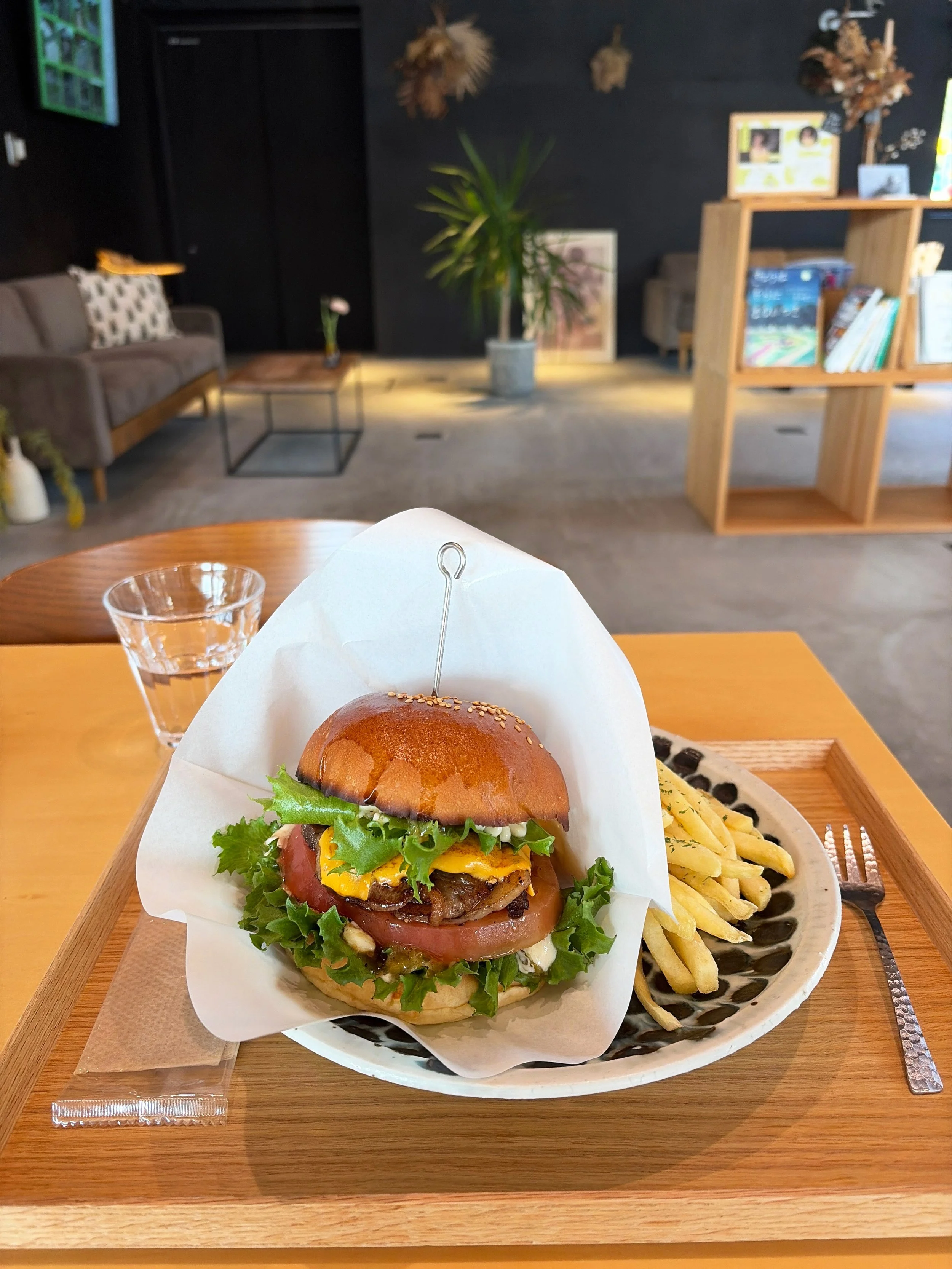 木目調のテーブルの上に、ボリュームのあるハンバーガーとフライドポテトが載っています。ハンバーガーは艶のある茶色いバンズに、ジューシーそうな肉、溶けた黄色いチーズ、赤いトマト、緑色のレタスが重なっています。白い紙で包まれ、中央には金属製のピックが刺さっています。奥にはグラスの水、ソファや観葉植物、本棚が見え、落ち着いたおしゃれな店内です。