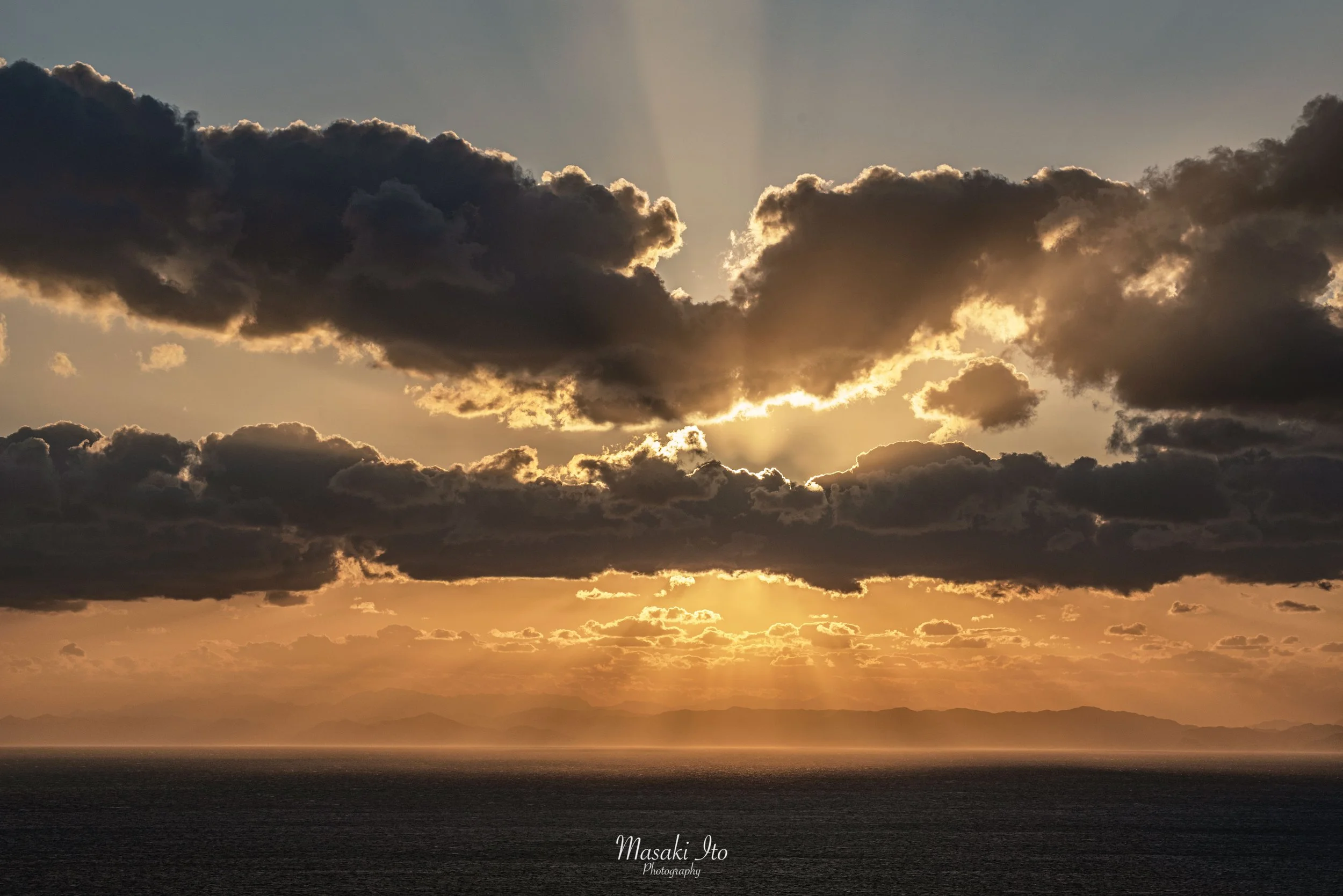 海の向こうに沈む夕日を、厚い雲が覆っている風景です。太陽は雲の後ろに隠れていますが、雲の隙間から黄金色の光が放射状に差し込み、雲の縁を金色に輝かせています。空は上部の暗い青灰色から、水平線に近づくにつれて鮮やかなオレンジ色へ変化。遠くには山々が霞んだシルエットとして浮かび、手前には静かな暗い海が広がっています。暗雲と夕日の光が対照的で、荘厳な雰囲気の一枚です。