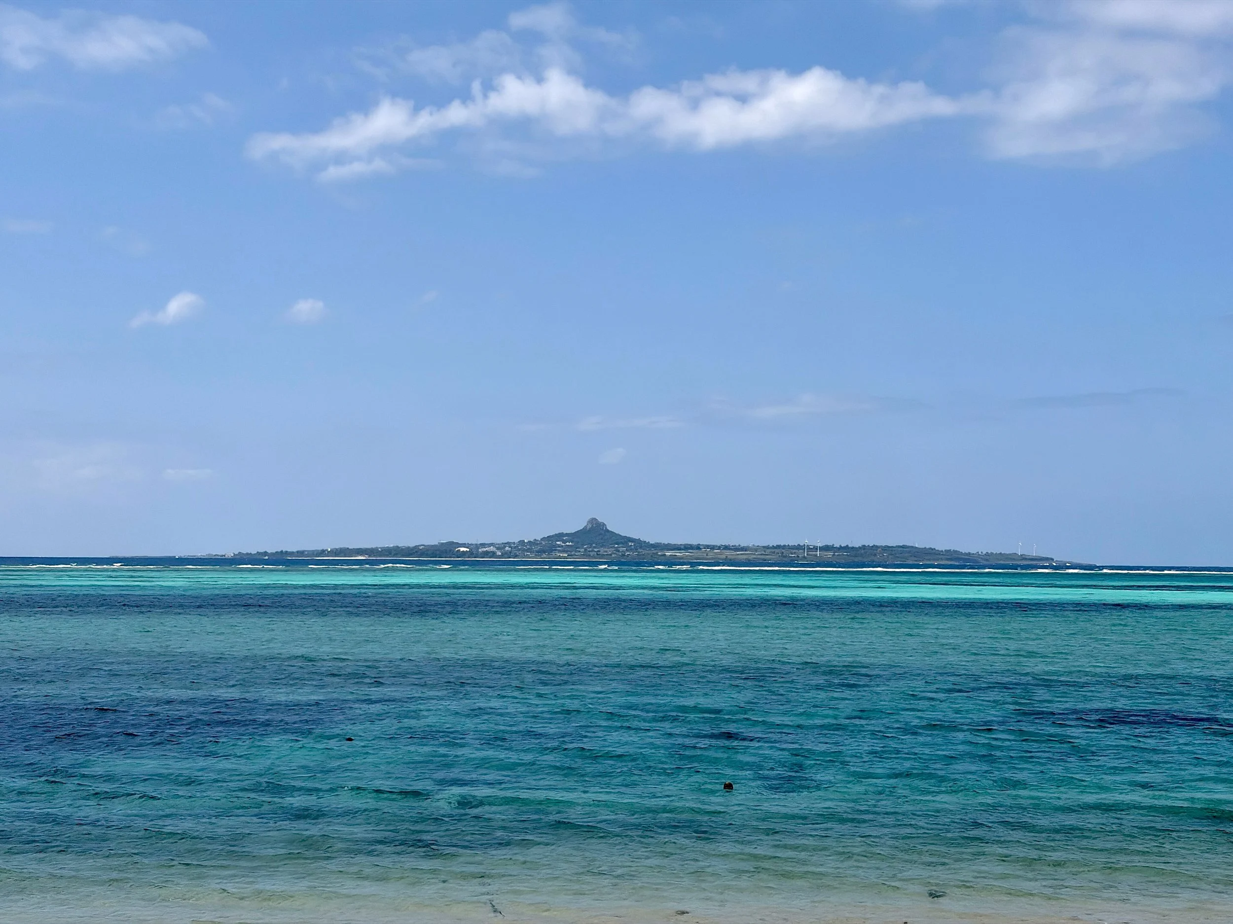 備瀬の浜から海を眺めた風景です。手前には、穏やかな波が広がる透明感のある青緑色の海があり、沖へ向かうにつれて濃い青色へと変化しています。水平線の向こうには細長い島影が浮かび、中央付近には特徴的な山が見えます。島の右側には小さな風力発電の風車も並んでいます。頭上には白い雲が浮かぶ、澄んだ青空が広がっています。