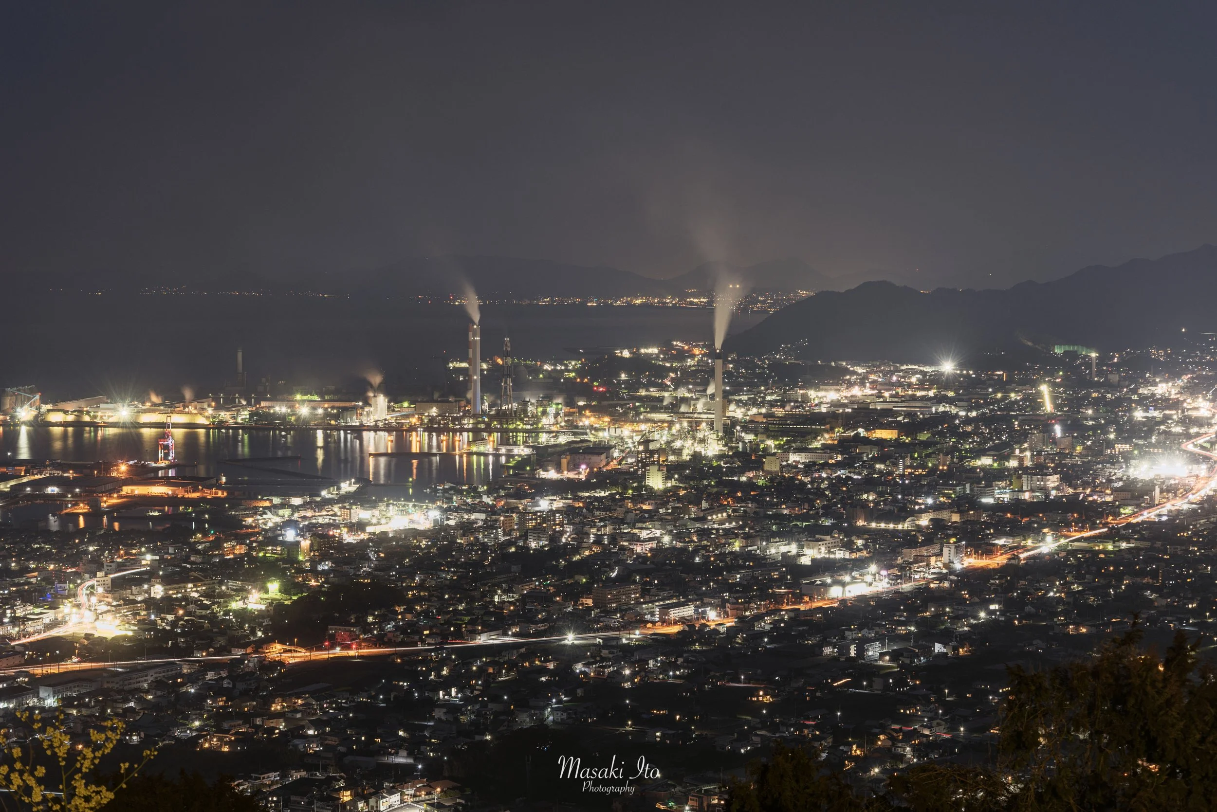 山の高台から見下ろした、愛媛県の夜景を捉えた写真です。眼下には住宅や建物が広範囲に広がり、無数の白やオレンジ色の明かりが街を彩っています。左側には大きな工場地帯と港があり、煙突から白い煙が立ち上っています。奥には海と暗い山々が連なり、手前から遠くまで街の光が続く、壮大な夜景です。