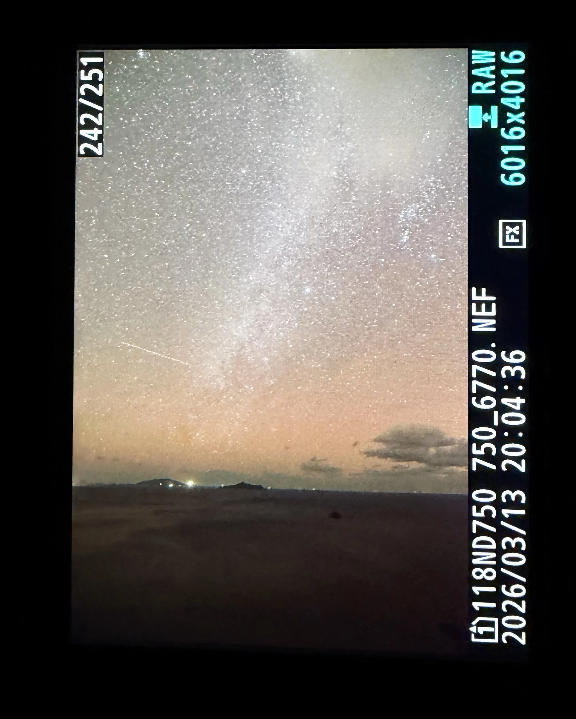 夜の海辺を写した写真です。空一面に無数の星が広がり、中央付近には淡く白い天の川が縦方向に伸びています。空の下部はオレンジ色に染まり、水平線近くには薄い雲が浮かんでいます。遠くには黒い島影が横たわり、いくつかの小さな明かりが輝いています。手前の海は暗く静かで、星空との境界が穏やかに広がる幻想的な夜景です。