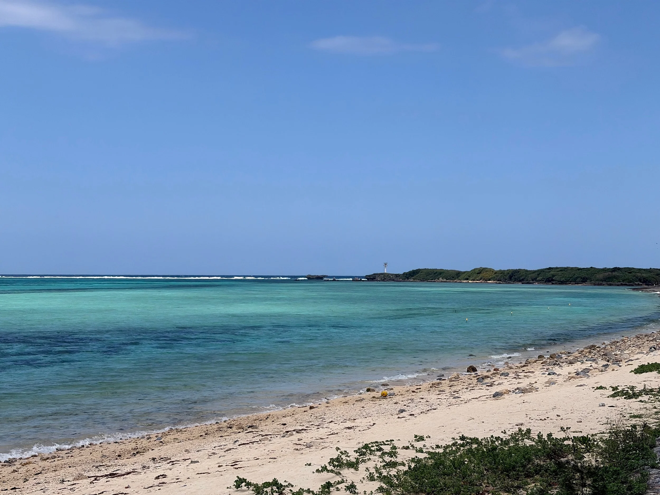 沖縄県本部町にある備瀬の浜を写した風景です。目の前には、透明感のあるエメラルドグリーンから青へと変化する穏やかな海が広がっています。沖合では白い波が一直線に立ち、遠くには緑に覆われた小さな岬と岩場、その先に灯台のような建物が見えます。手前には白い砂浜が緩やかに続き、大小の石が散らばっています。晴れた青空と海の色が美しく、南国らしい開放感を感じる一枚です。