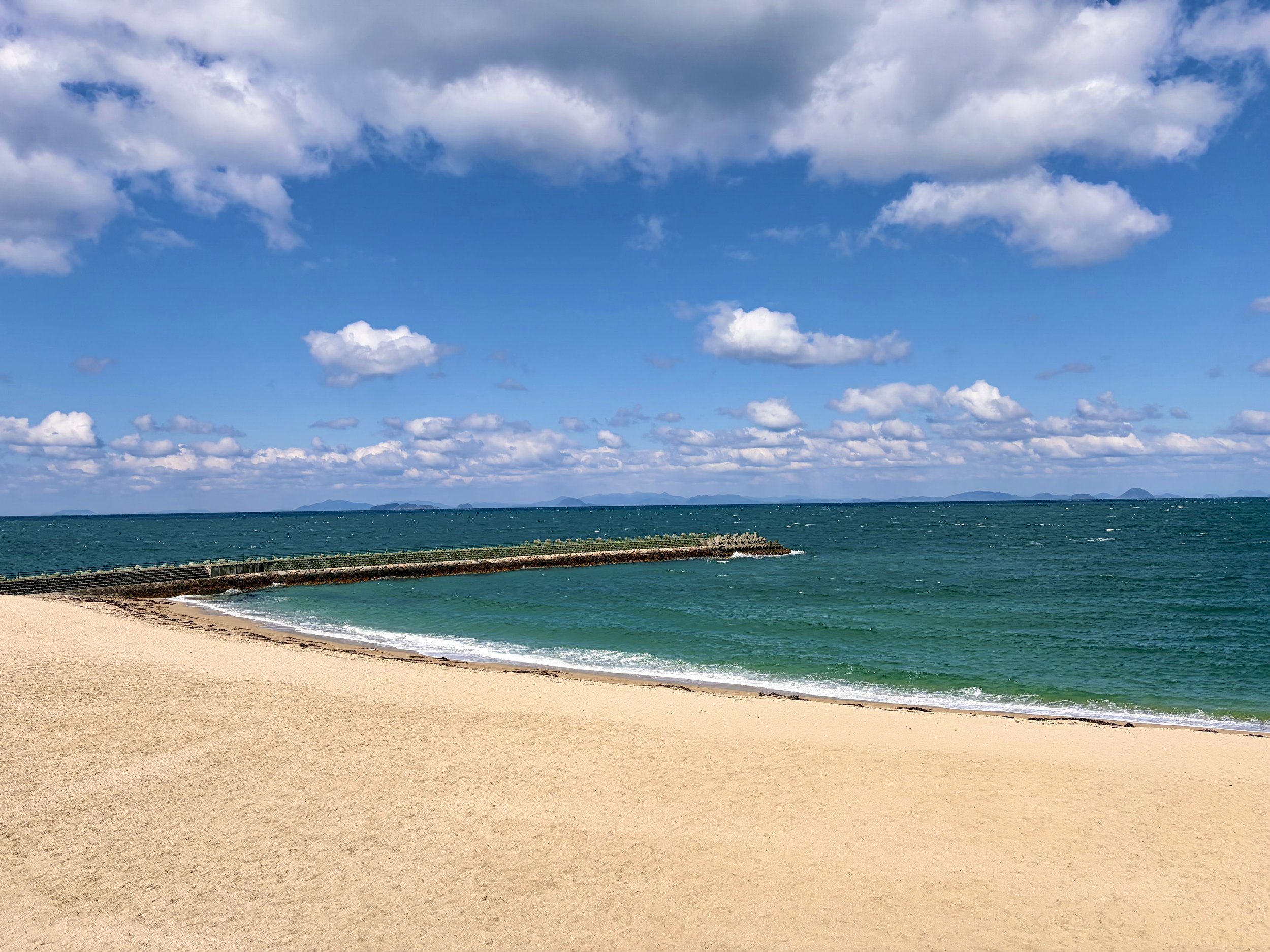 広い砂浜と青い海が広がる、開放感のある海岸風景です。手前には淡いベージュ色の砂浜が一面に広がり、その先で白い波が静かに打ち寄せています。海には細長い防波堤が伸び、遠くの水平線には小さな島々や山々が連なっています。空は鮮やかな青色で、大小の白い雲が浮かび、海と空の青さが美しく調和しています。