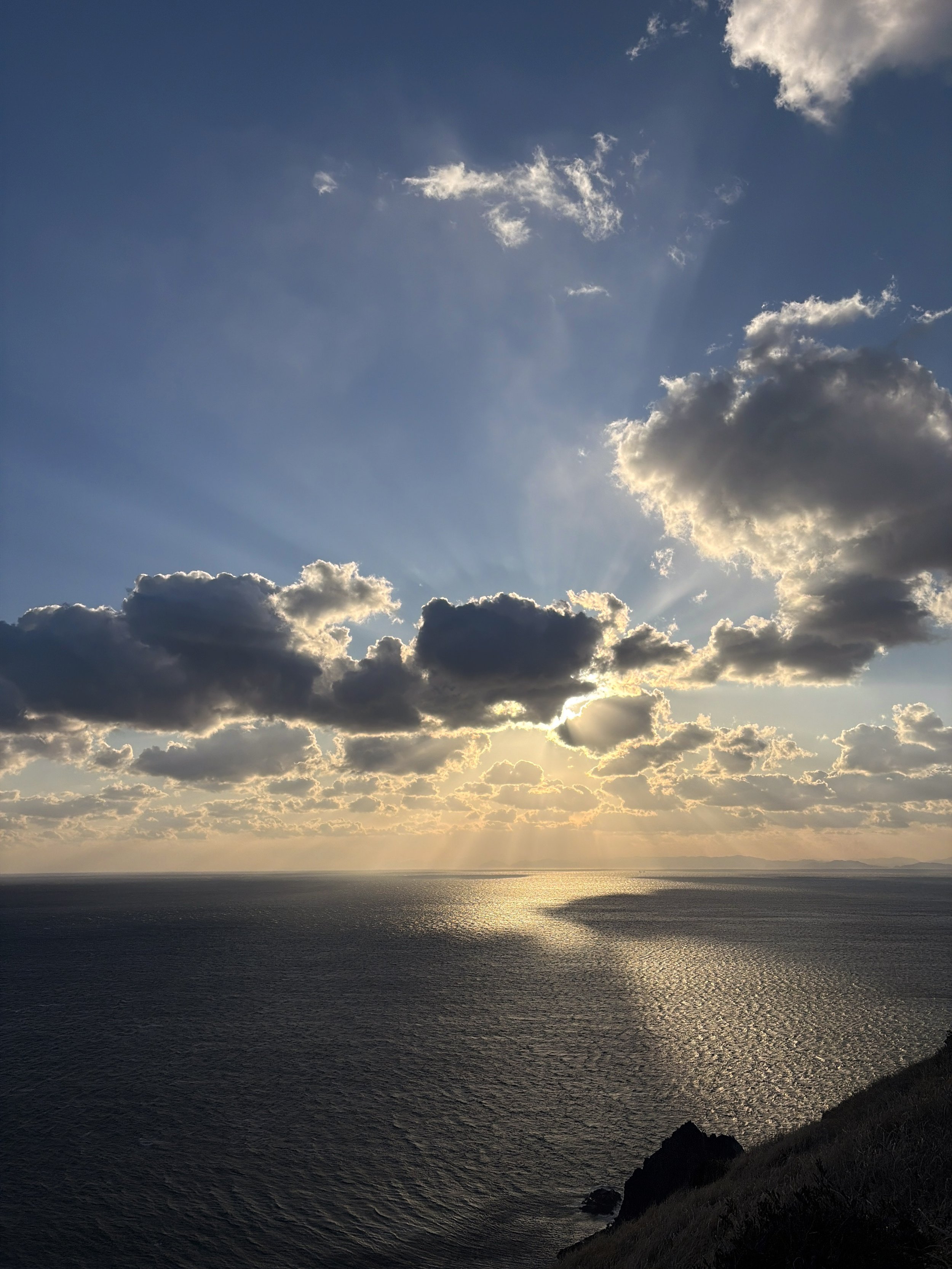 高台から広大な海と空を見渡した夕暮れの風景です。青い空には大小の雲が浮かび、雲の後ろに隠れた太陽から何本もの光が放射状に伸びています。黄金色の光は海面にも反射し、水平線から手前へ一本の輝く道を描いています。右下には暗い岩場と斜面があり、青い海と金色の光の対比が、雄大で神秘的な雰囲気を感じさせます。