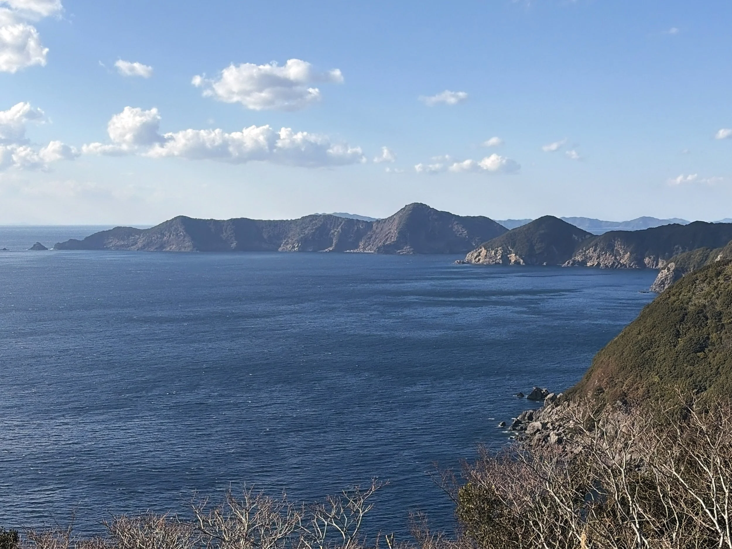 青い海を見下ろす高台から、対岸の山々を望んだ風景です。穏やかな海が画面いっぱいに広がり、遠くには複雑な形の山々が連なっています。山の斜面には緑の木々が広がり、海岸には白っぽい岩場が見えます。澄んだ青空には小さな白い雲が浮かび、海と山、空が美しく調和した開放感のある景色です。