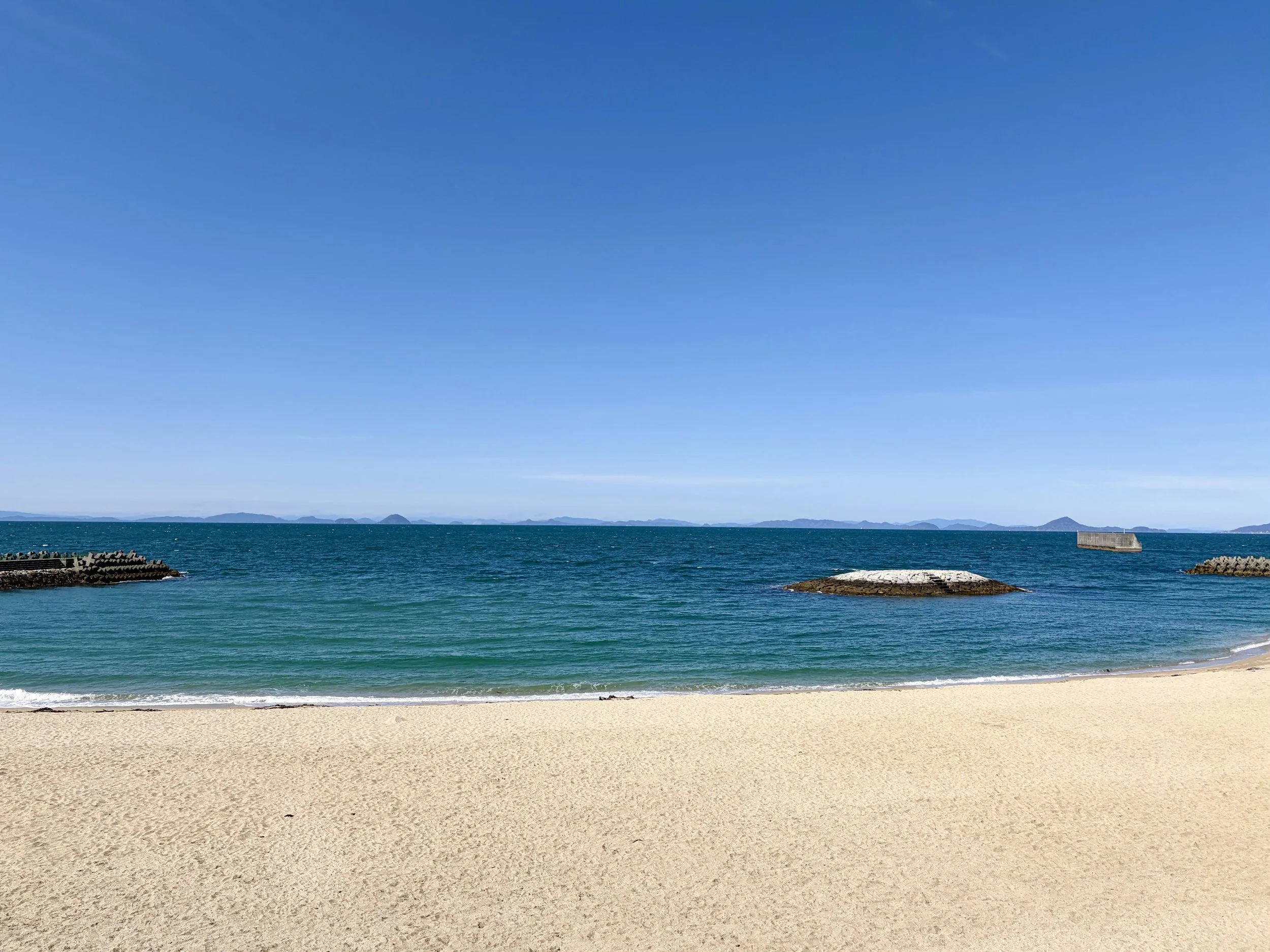目の前に広い砂浜があり、きめ細かな淡い色の砂が一面に広がっています。その先には青く透き通った海が広がり、穏やかな波が砂浜へ静かに打ち寄せています。海の上には小さな島や防波堤が点在し、遠くにはいくつもの島影が水平線に連なっています。空は雲がほとんどない澄んだ青空で、海と空がつながるような開放感のある美しい海辺の景色です。