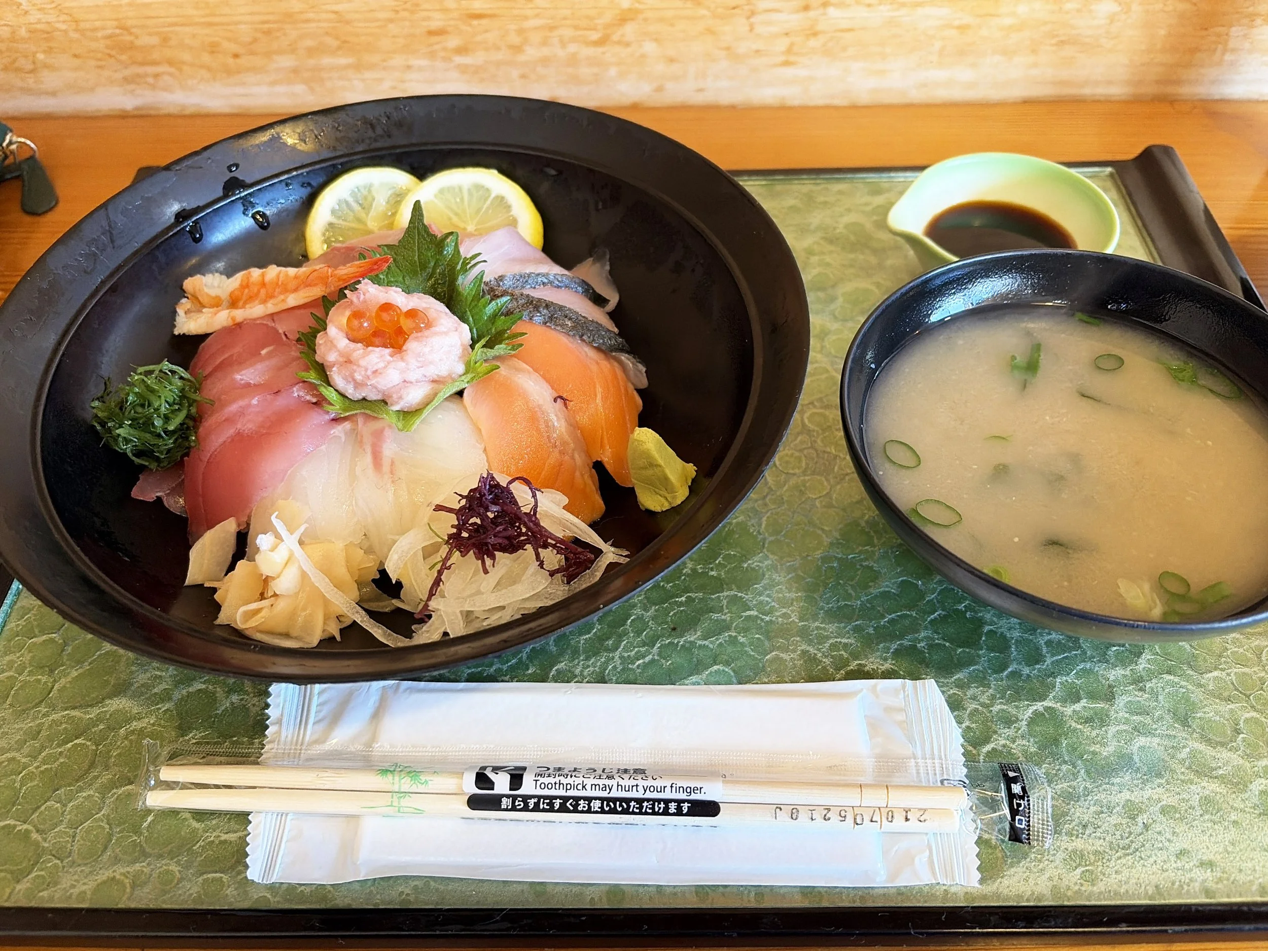 黒い器に盛られた海鮮丼と味噌汁の定食です。海鮮丼には、赤身のマグロ、白身魚、サーモン、エビなど色とりどりの刺身が並び、中央にはイクラを添えたネギトロが盛られています。レモンや大葉、ワサビ、ガリも添えられています。右にはネギの浮かぶ味噌汁、奥には醤油皿があり、手前には割り箸が置かれています。