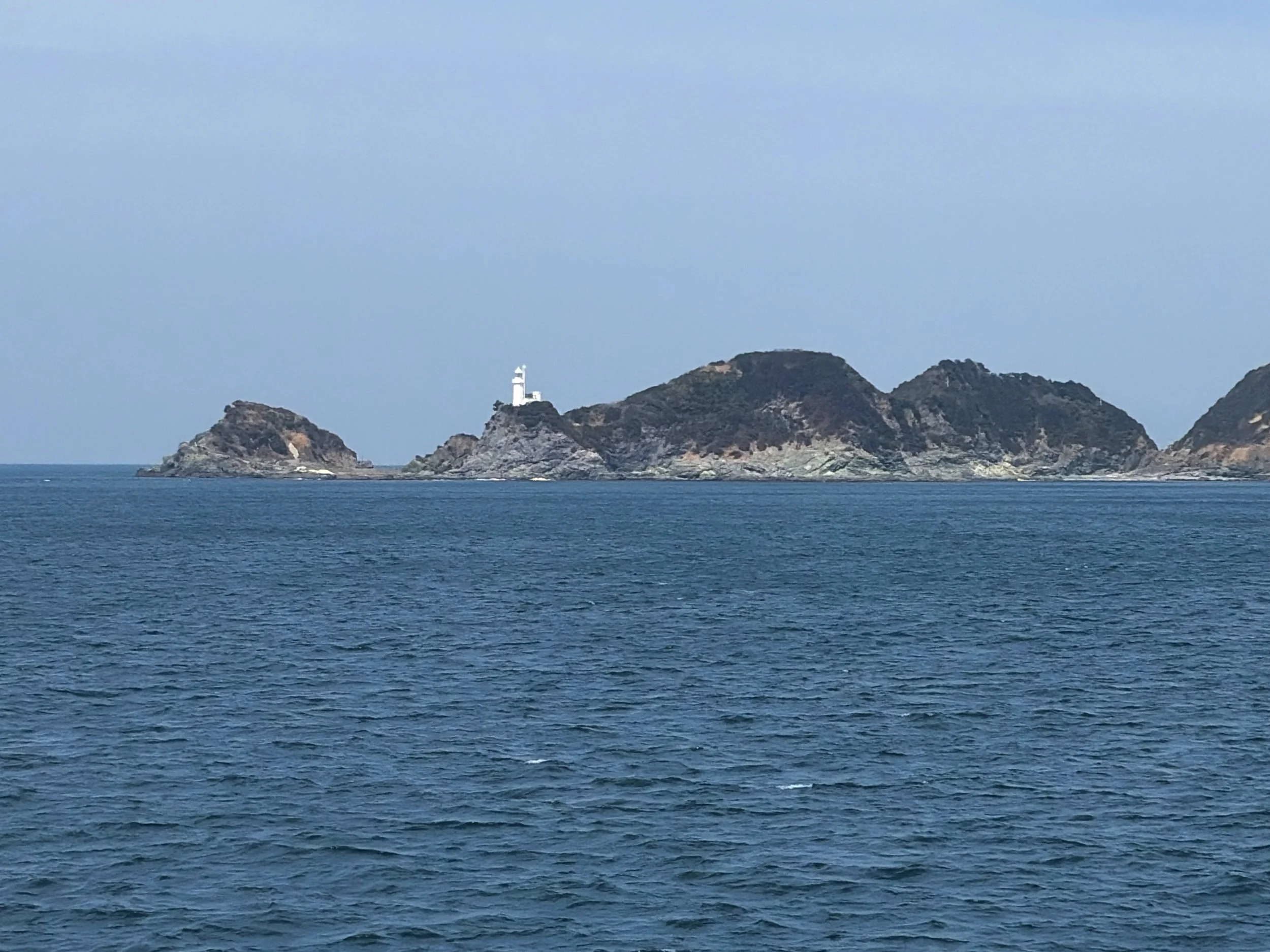 海の向こうに、いくつもの岩礁の島が横一列に連なっています。手前には濃い青色の海が広がり、細かな波が静かに揺れています。中央付近の岩の上には、小さな白い灯台が立っています。島々は黒や灰色の岩肌が目立ち、ところどころ茶色の部分もあります。背景は雲の少ない淡い青空で、海と島の境界がくっきりと見える、穏やかな海の風景です。