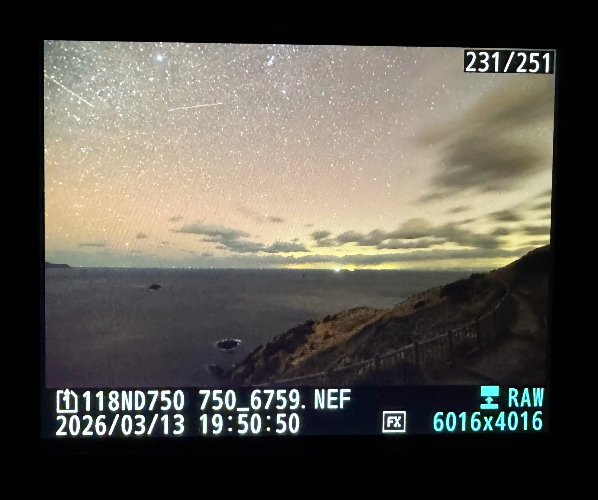 夜の海を見下ろす高台から撮影した風景です。空には無数の星が広がり、薄く雲が流れています。水平線付近には黄色や白色の光が帯状に輝き、海と空の境界を照らしています。右側には斜面と海沿いへ続く細い遊歩道が見え、手前から遠くへ伸びています。静かな夜の海と星空、海岸線の光が幻想的に重なった一枚です。