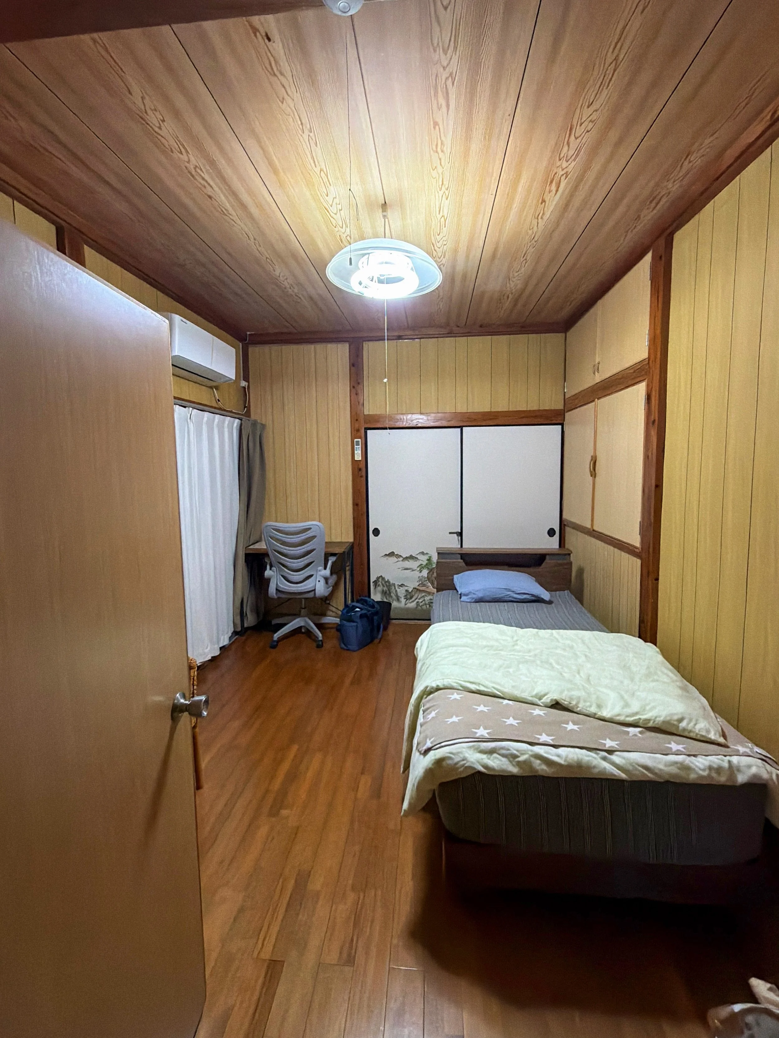 木の温もりを感じる、和風の落ち着いた一人用の部屋です。天井と壁は木板で覆われ、床も木目調で統一されています。右側には一台のベッドがあり、薄い緑色の掛け布団と星柄の毛布、青い枕が置かれています。正面には山水画が描かれた白い襖があり、その手前には小さな机があります。左側にはカーテン付きの窓とエアコン、椅子、旅行用のバッグがあります。天井中央の照明が部屋全体を明るく照らしています。