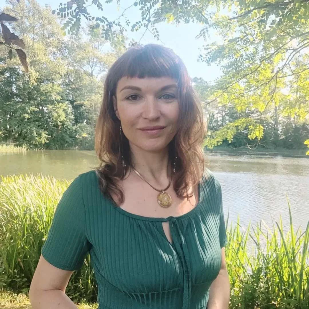 Eine Frau mit braunen, lockigen Haaren und Flügelohrringen steht an einem Fluss im Grünen, während die Sonne durch die Blätter scheint.