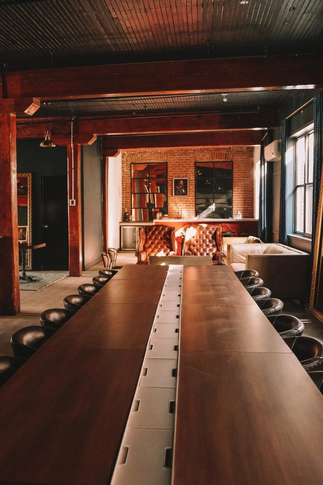 A large wooden conference table with black leather chairs, in an industrial-style room with brick walls, dark wood beams, and large windows, decorated with vintage portraits and furniture.