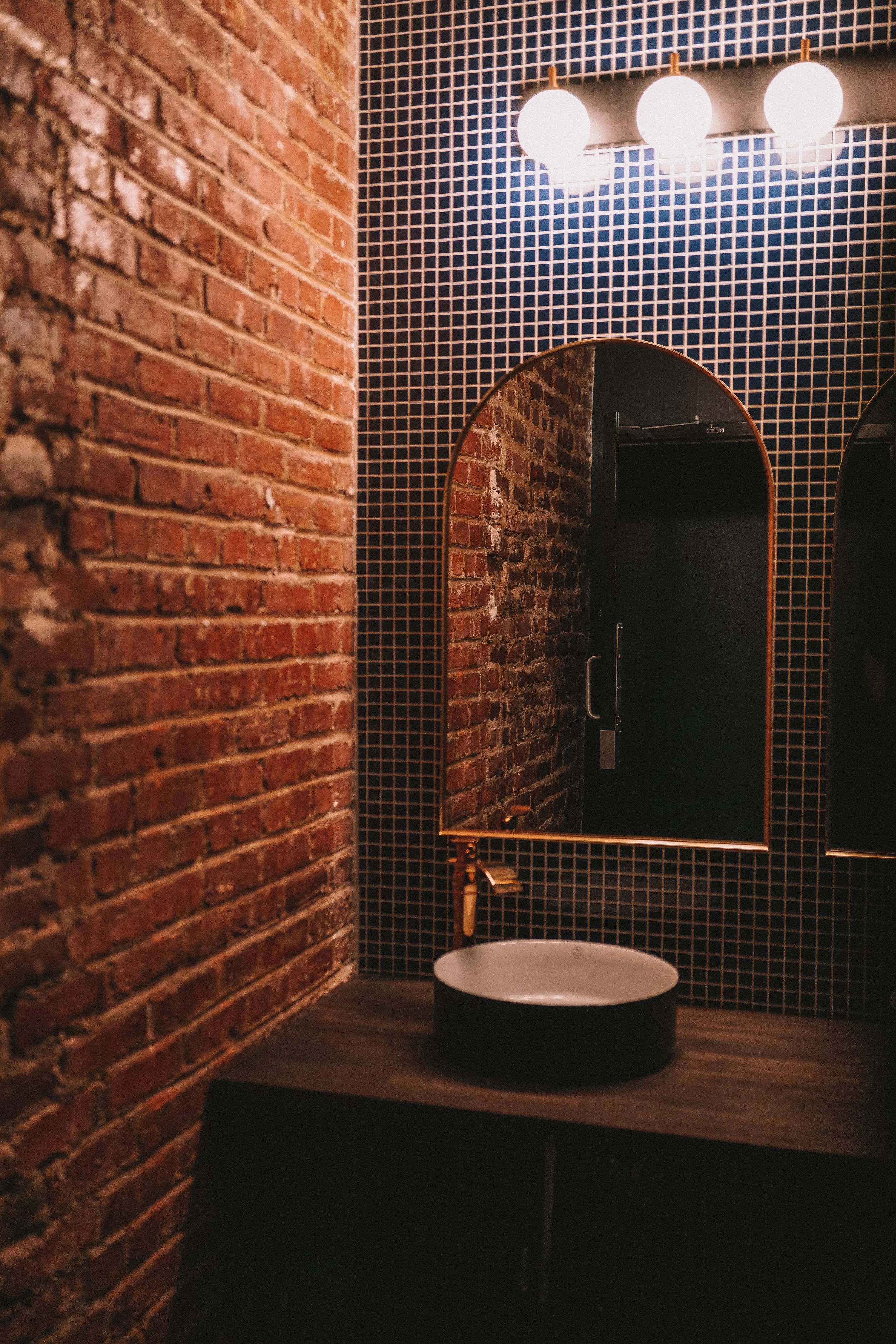 Industrial-style bathroom with a brick wall, a round vessel sink on a dark wooden countertop, a black-tiled wall with an arched mirror, a brass faucet, and three globe light fixtures above.