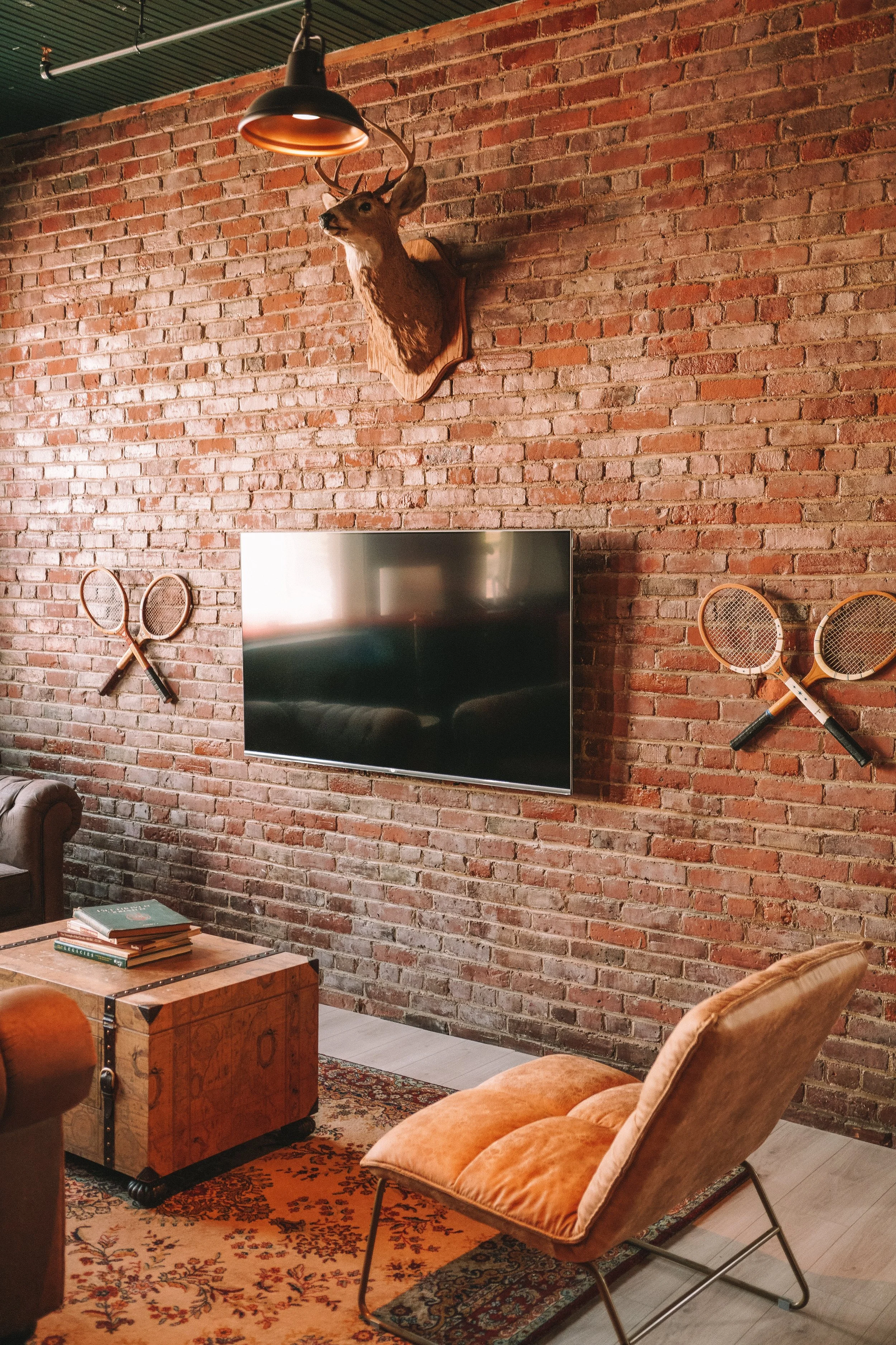 Interior of a room with a brick wall, a mounted deer head, a television, vintage tennis rackets on the wall, and orange velvet chairs. A wooden coffee table with books is also visible.