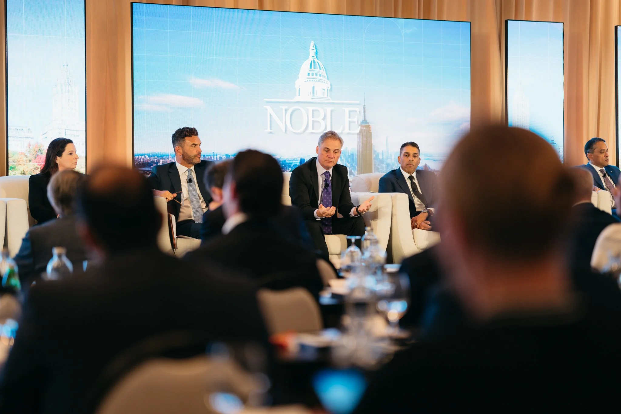 Panel discussion at a conference with a large screen displaying the word NOBLE and an illustration of a government building.