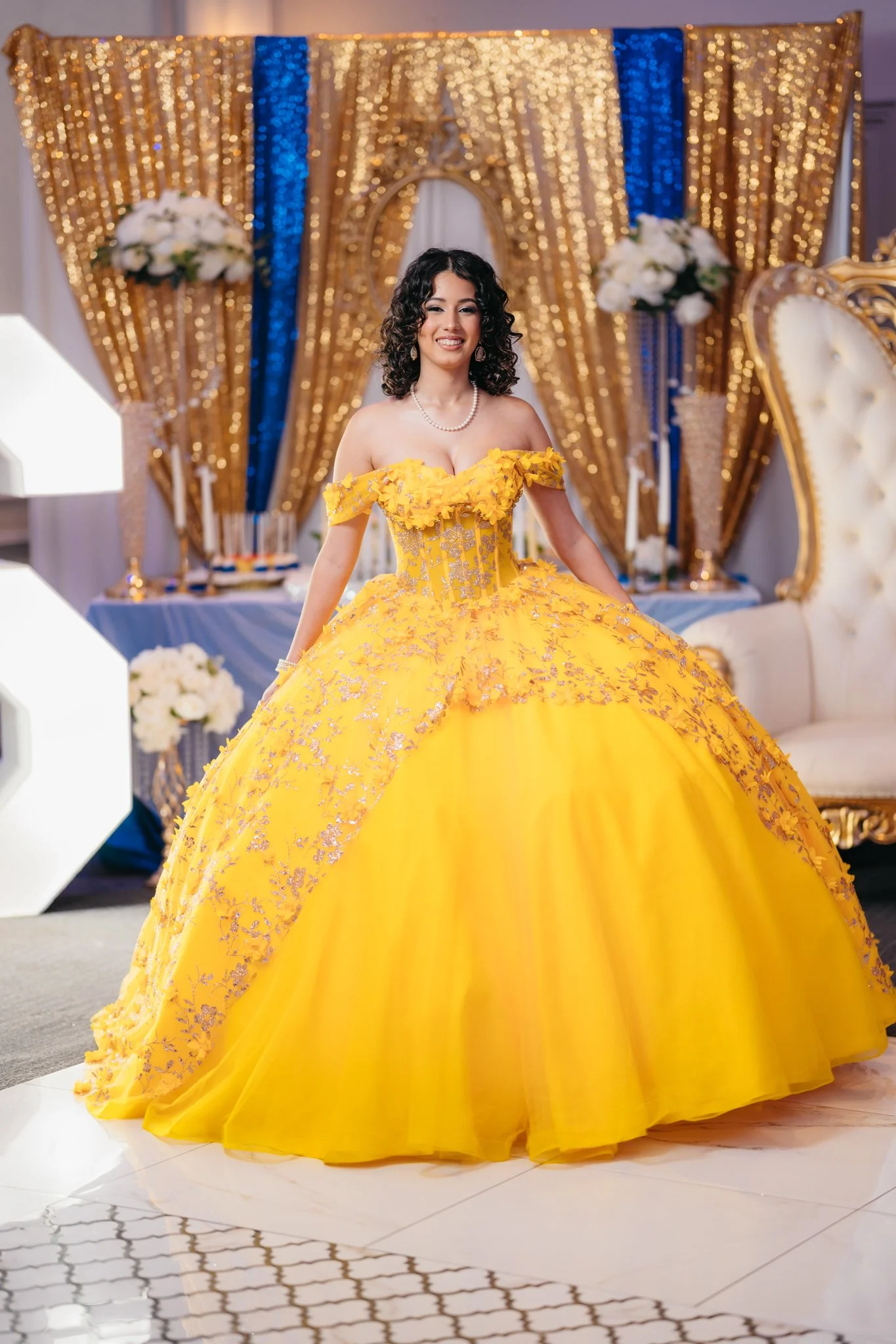 A woman in a yellow quinceañera dress, decorated with floral appliques, standing in a decorated event space with gold and blue curtains, white floral arrangements, and ornate furniture.
