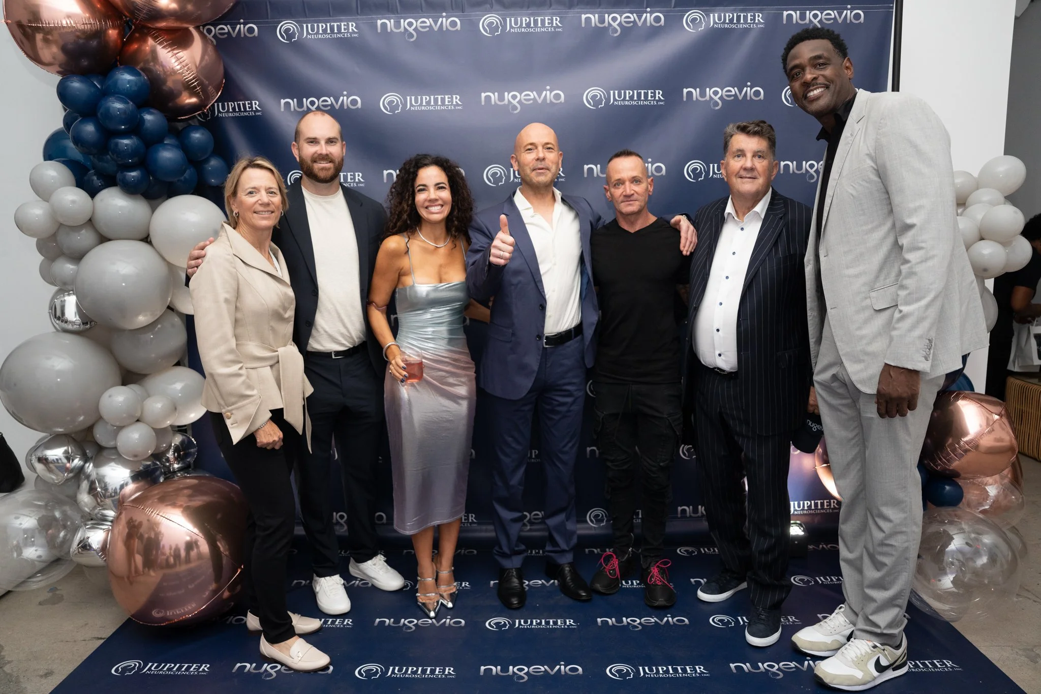 A group of eight diverse people posing and smiling at a celebration event with a backdrop that has 'Jupiter Neurosciences, Inc.' and 'Nugevia' written on it, standing on a black carpet surrounded by balloons.