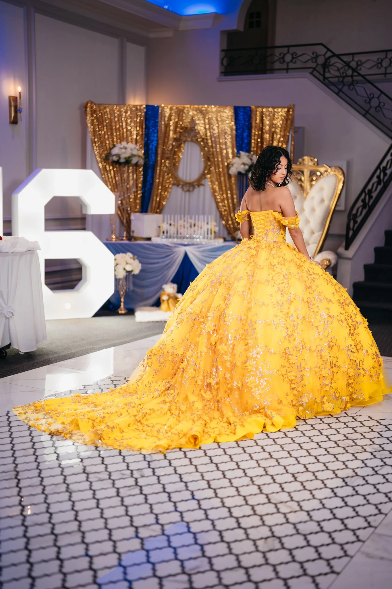 A woman in a bright yellow ball gown with floral embroidery stands in an elegant decorated venue. The background features a gold and blue curtain, floral arrangements, and a white decorative letter 'Q'.