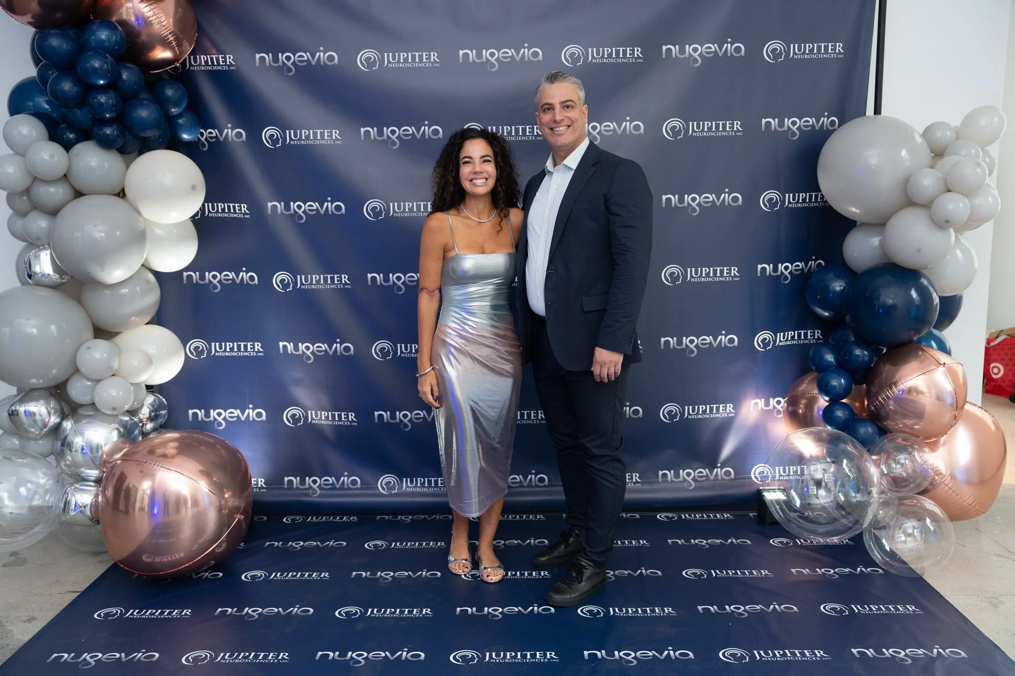 A woman and a man pose together at a professional event in front of a blue backdrop with logos of Noveva and Jupiter Neurosciences. The woman is wearing a shiny, metallic silver dress and heels, and the man is in a dark suit with a white shirt. They 