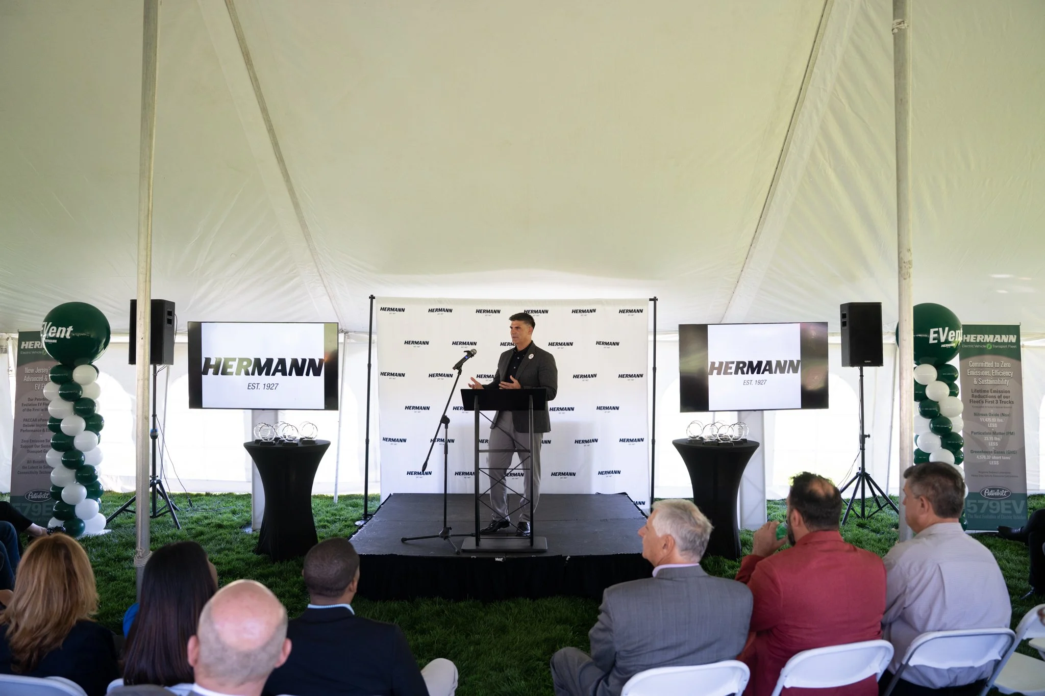 A man in a dark suit speaking at a podium on a stage inside a large white tent. Behind him are two screens displaying the Hermann logo and setup tables with awards. Audience members are seated facing the stage, with some taking notes or listening att
