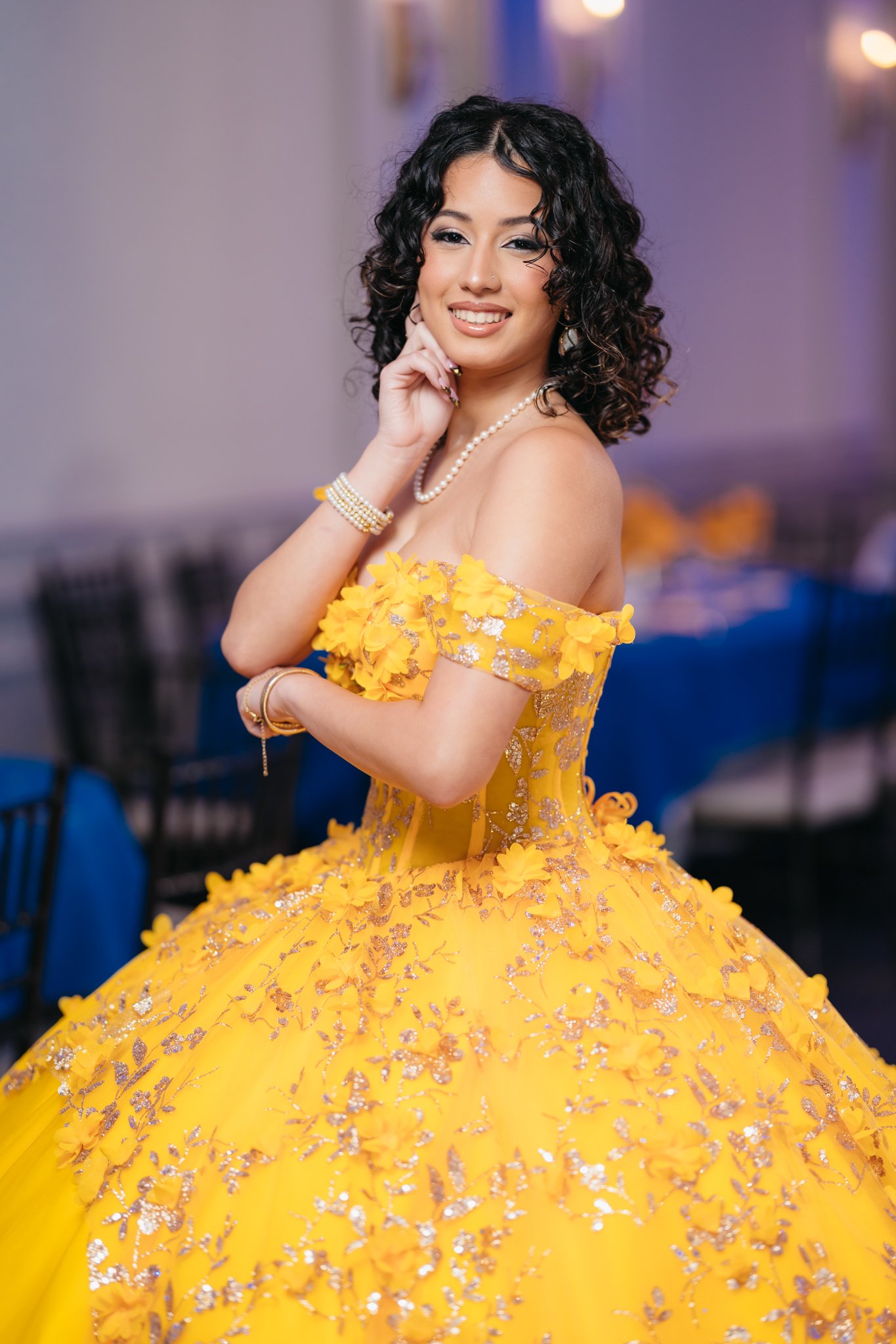A young woman dressed in a yellow floral ball gown with off-shoulder sleeves, accessorized with pearl jewelry, standing indoors at a formal event.