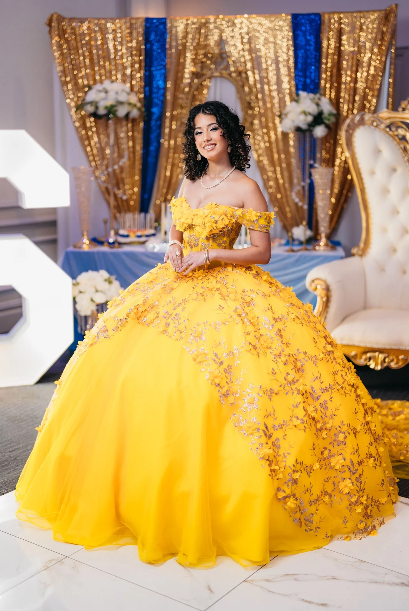 A young woman in a vibrant yellow ball gown with floral embroidery, smiling at a celebration event.