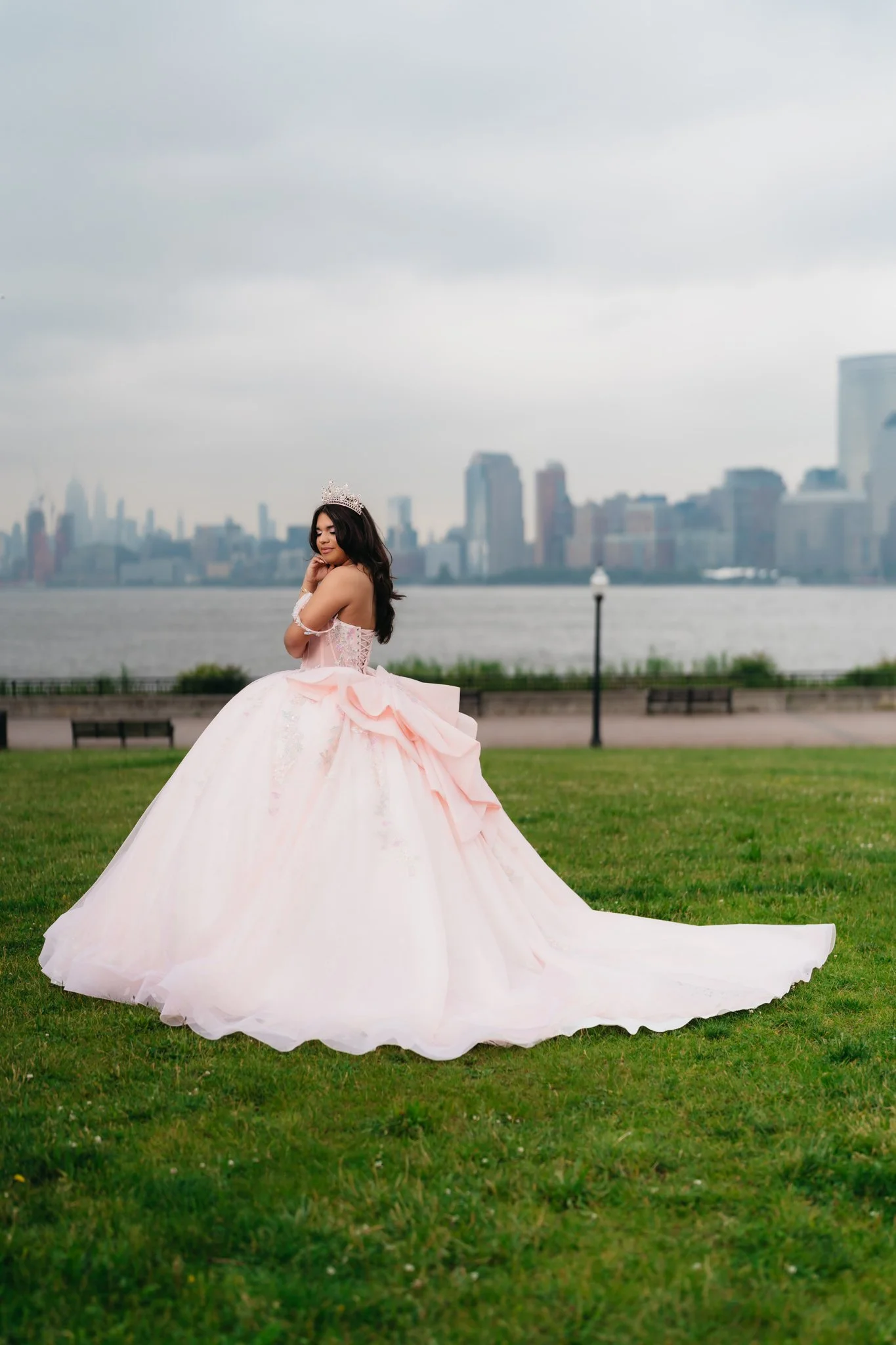 Young woman in a pink ball gown and tiara standing on grass with city skyline in the background.