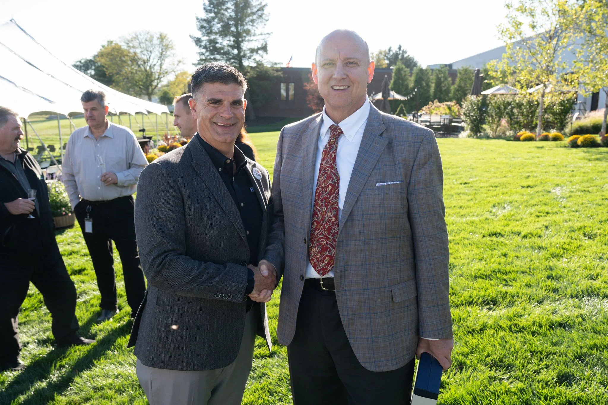 Two men in suits shaking hands outdoors on a sunny day, with other people and green grass in the background.