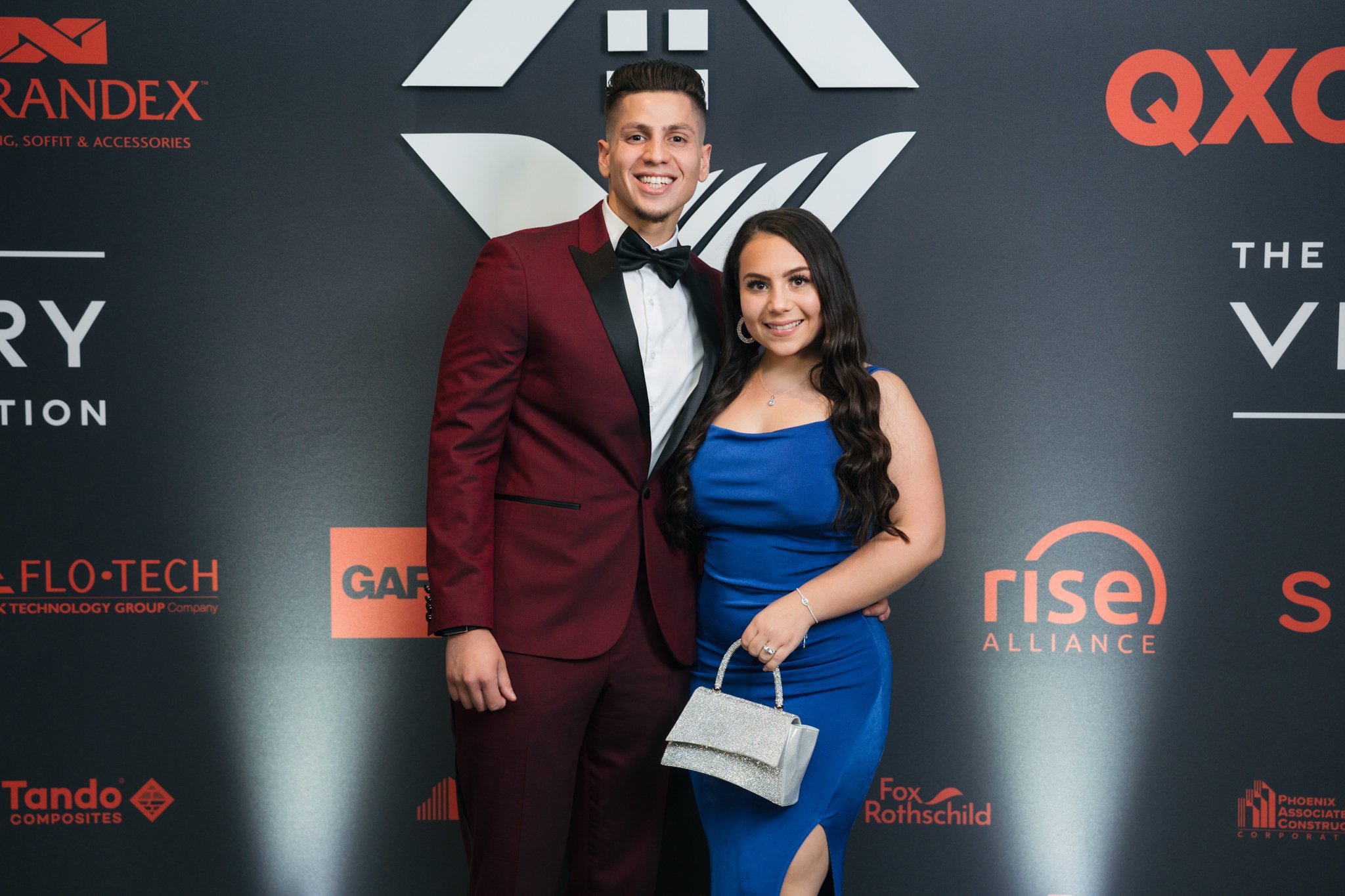 A young man and woman dressed in formal attire posing together at an event with a dark background and various sponsor logos.