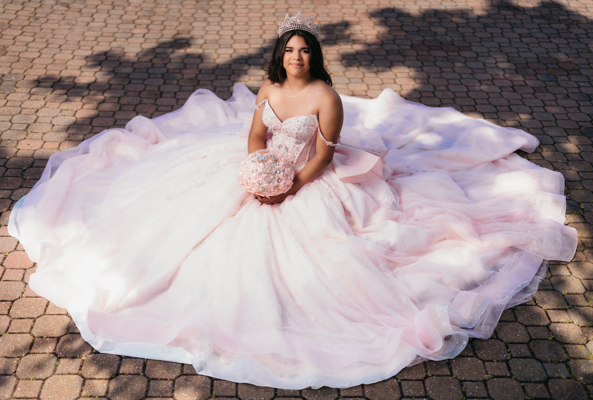 Young woman in a pink ball gown sitting on brick pavement, wearing a tiara, holding a bouquet of pink roses.