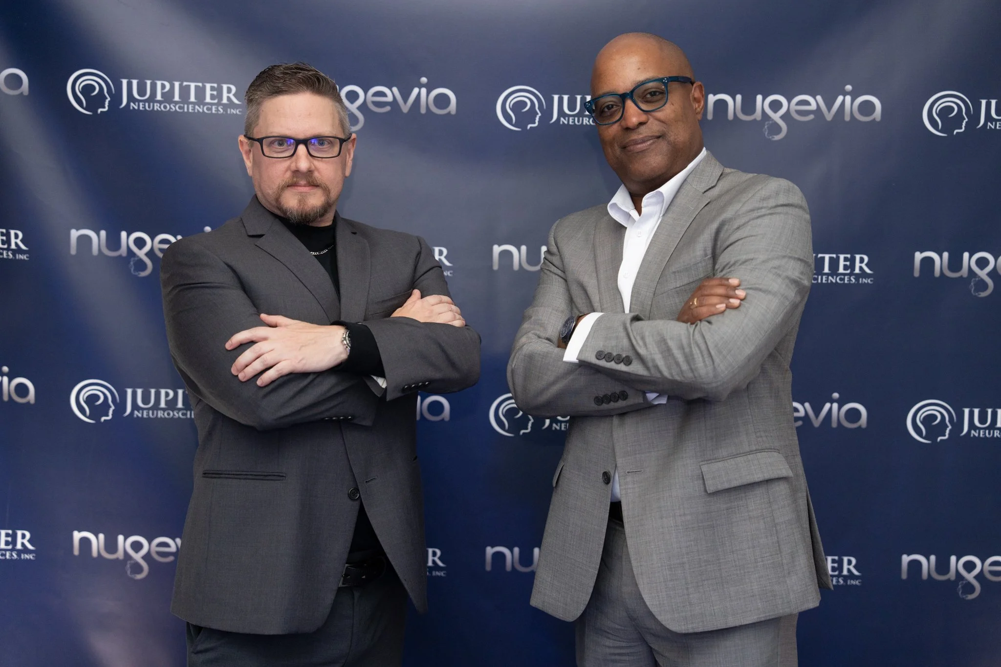 Two men in business suits standing with arms crossed in front of a blue backdrop with logos for JUPITER NEUROSCIENCES, Inc., and nuveia.