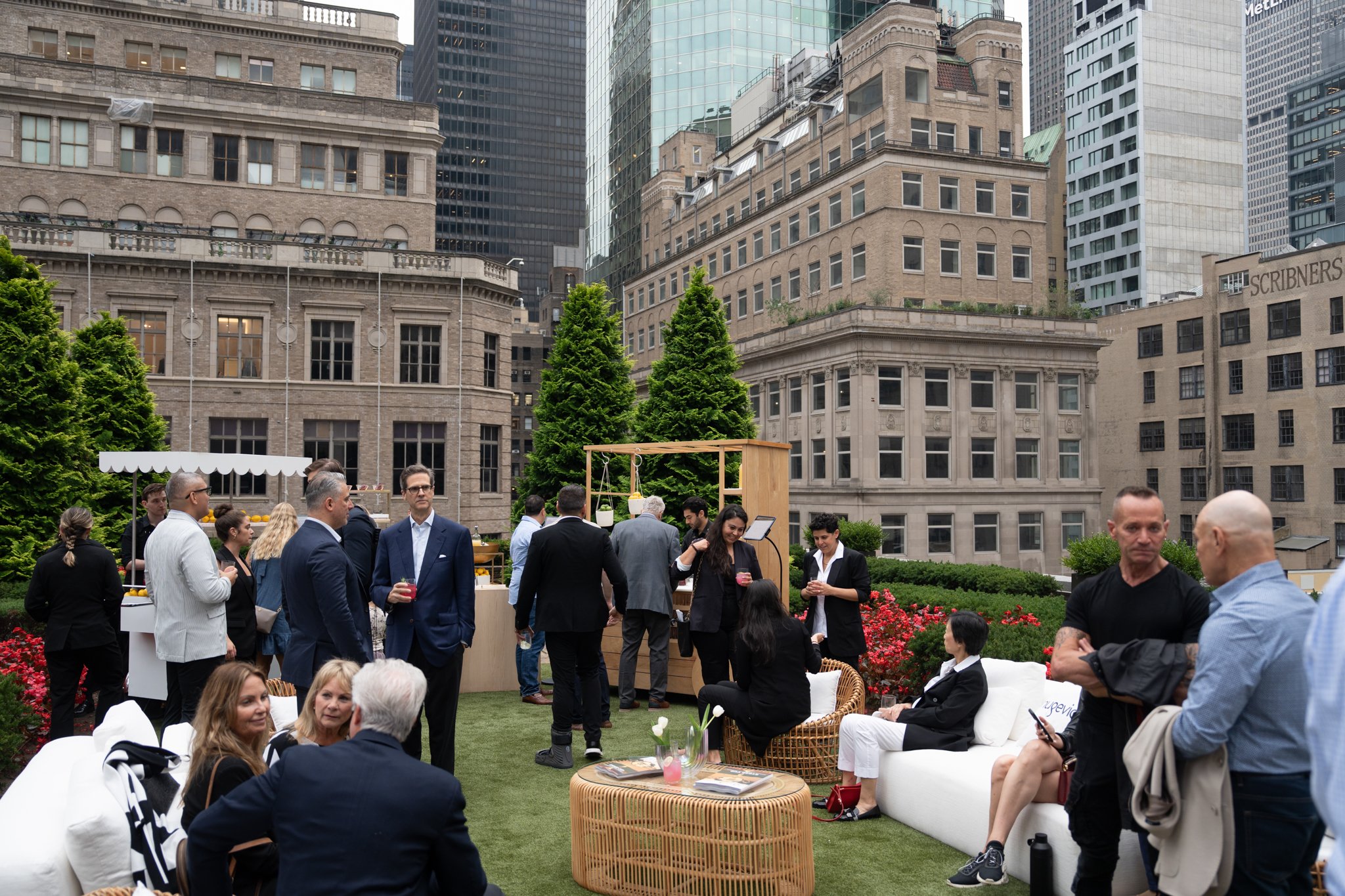People enjoying a rooftop social gathering in an urban setting with high-rise buildings, sitting on outdoor furniture, chatting, and standing around a bar.