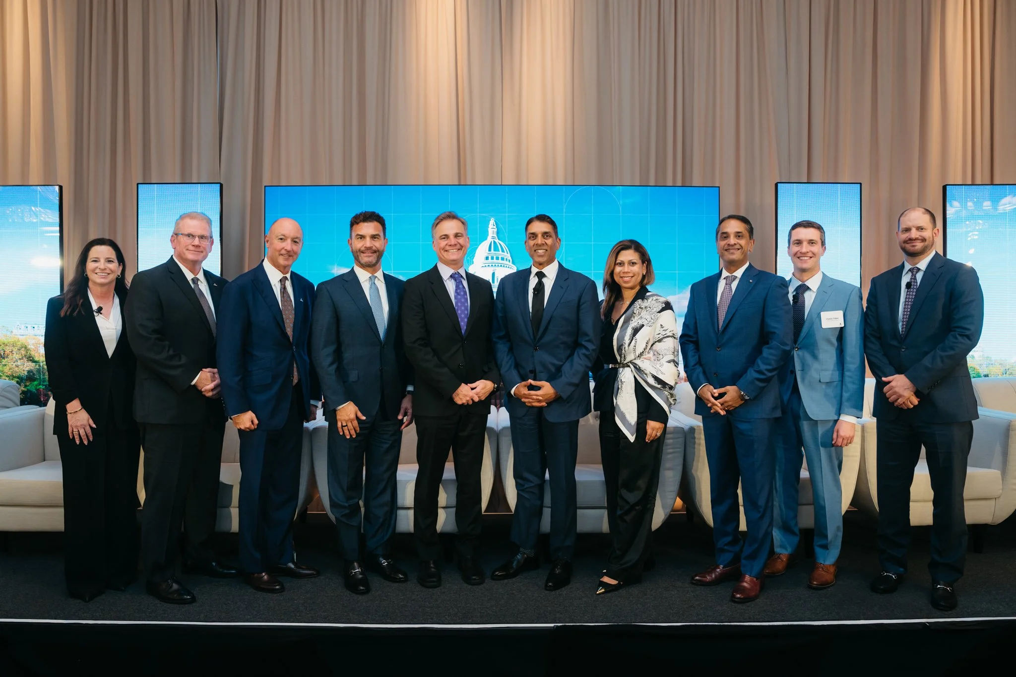 Group of eleven professionals in business attire standing on stage in front of a large blue digital display with an image of the Capitol building, in a formal conference setting.