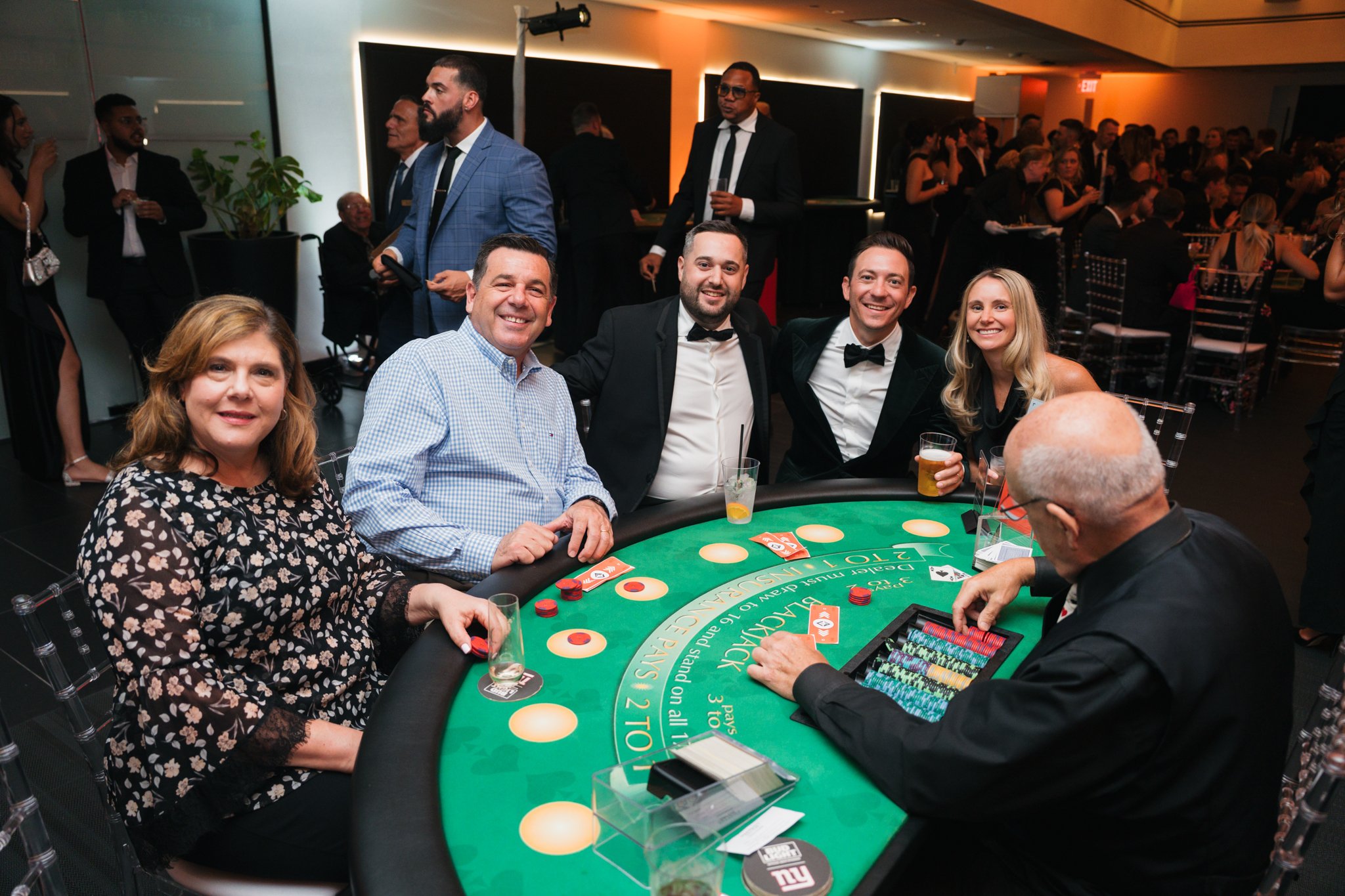 Group of people at a casino, sitting around a blackjack table, dressed in formal attire, with some holding drinks and others playing the game.