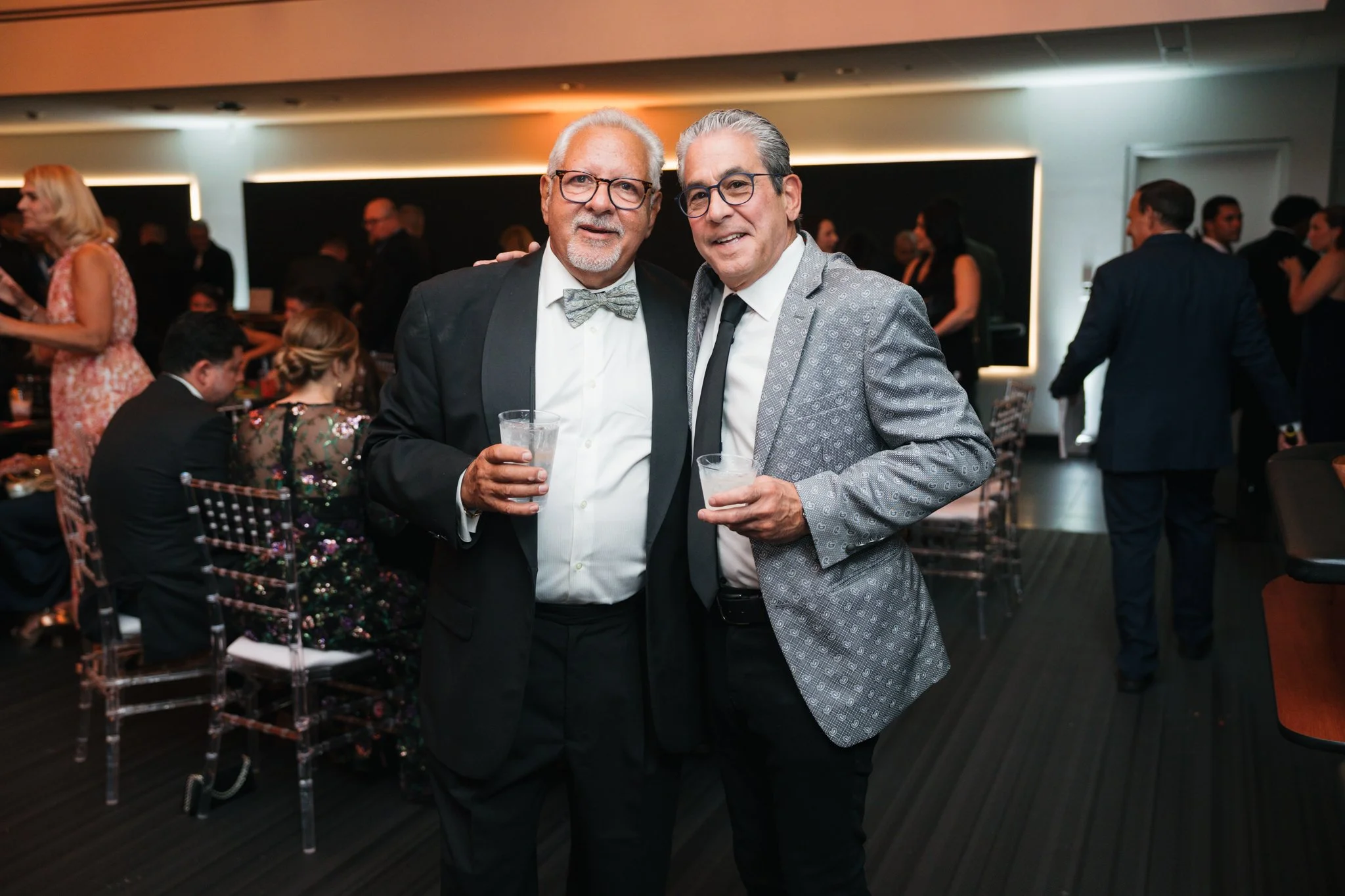 Two men in formal attire holding drinks at a social event or party, with other guests in the background.