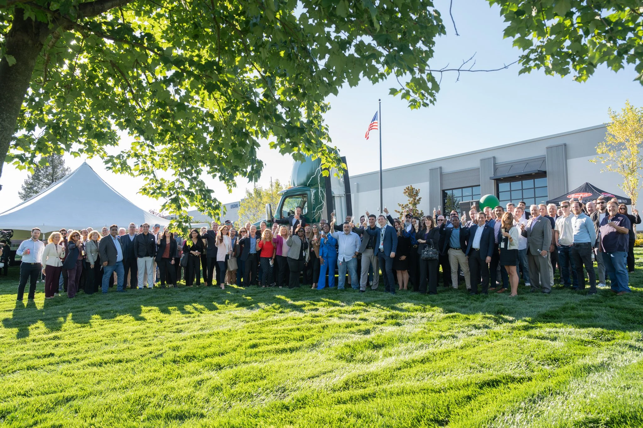 A large group of people standing outdoors on grass in front of a building with a truck parked behind them, trees, and a tent to the left. The group is diverse and dressed in business attire, celebrating at an event on a sunny day with some people hol