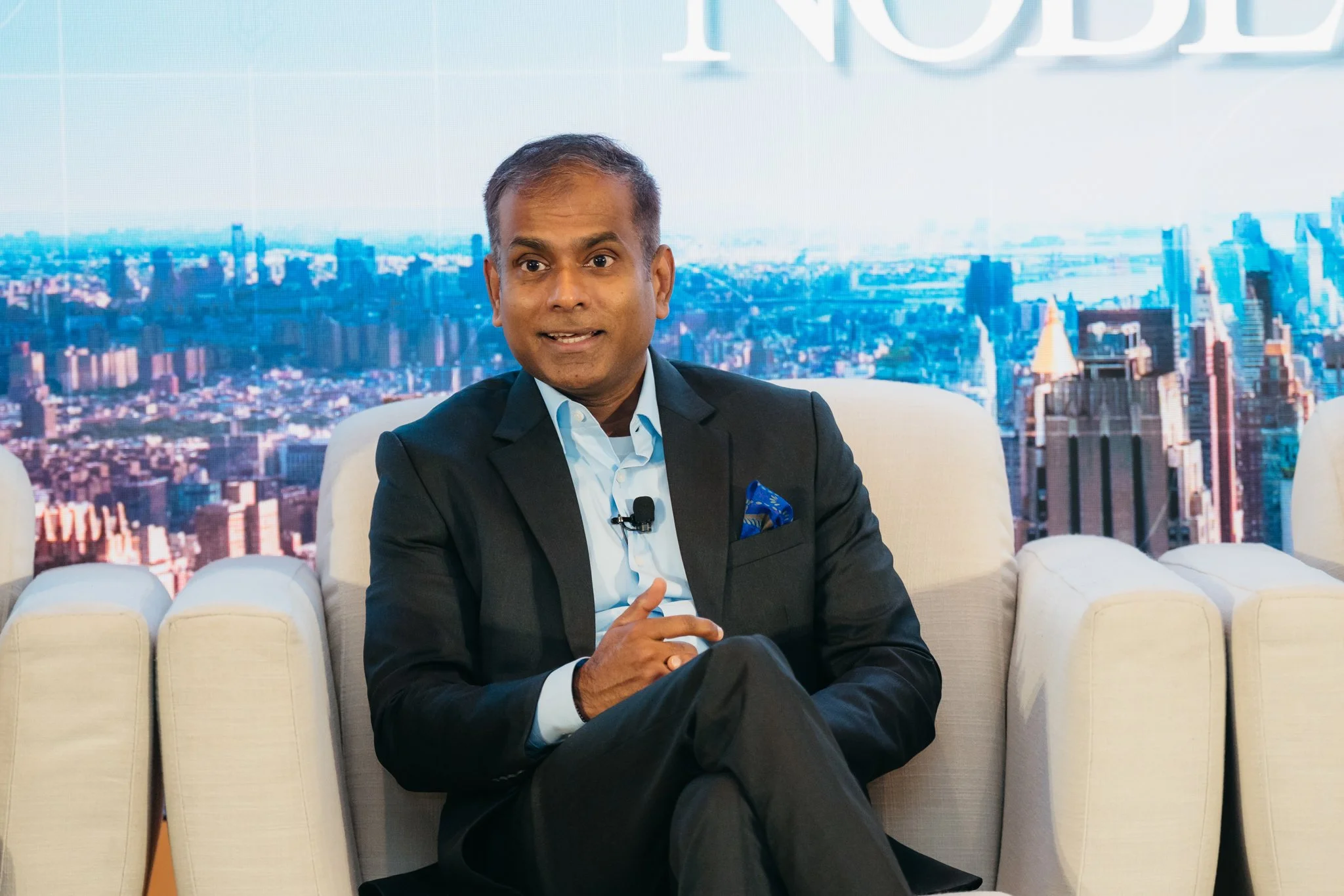 A man in a black suit and white shirt sitting on a beige sofa, speaking during a panel or interview, with a cityscape backdrop featuring tall buildings and a partial word 'NOD' visible in the background.