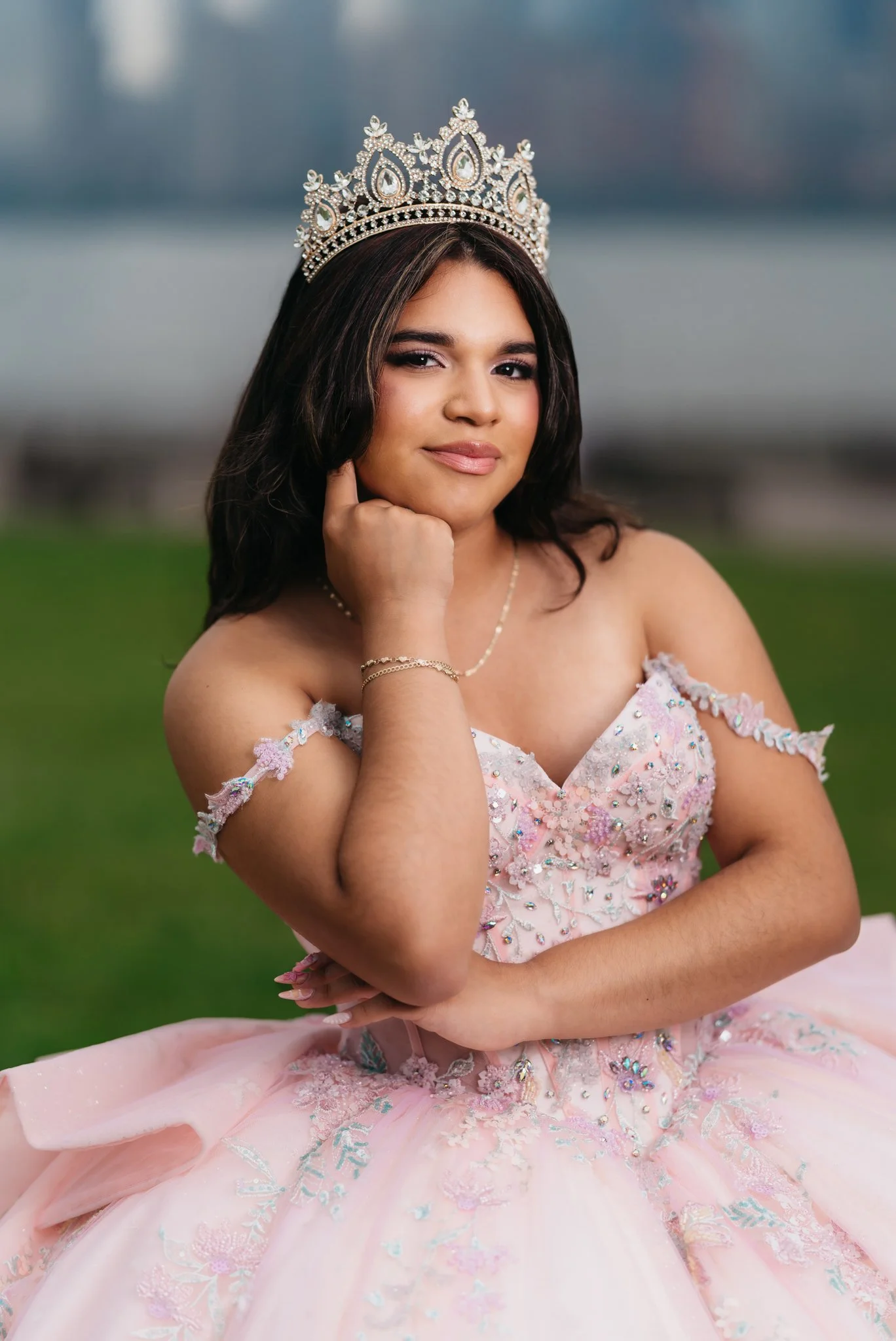 Young woman wearing a pink embellished gown and a jeweled crown, sitting outdoors with a blurred background.