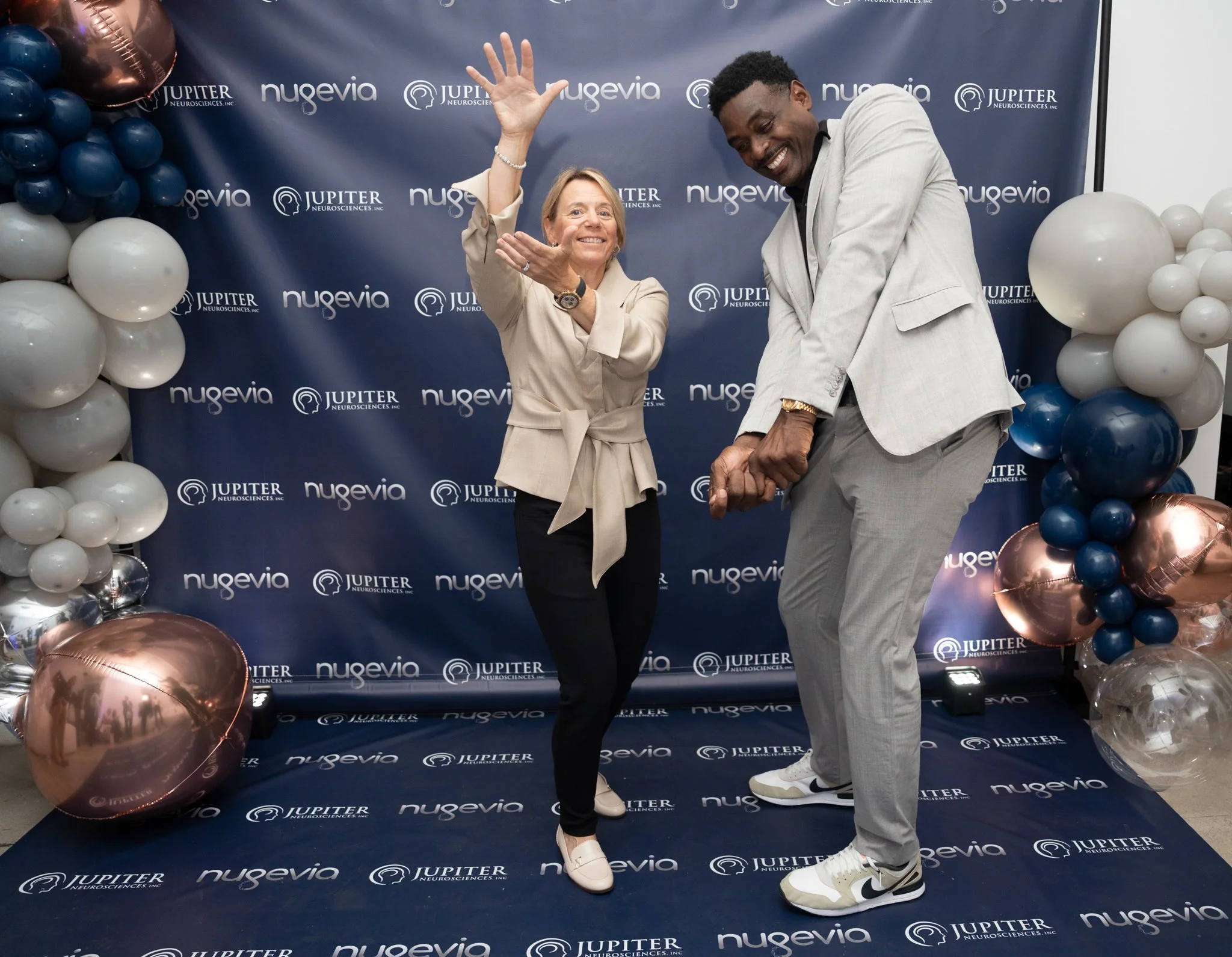 Two people dancing and smiling in front of a blue backdrop with the logos of Nugevia and Jupiter Neurosciences. The woman is raising one arm and wearing a beige blazer, black pants, and white shoes. The man is in a gray suit and sneakers, holding a d
