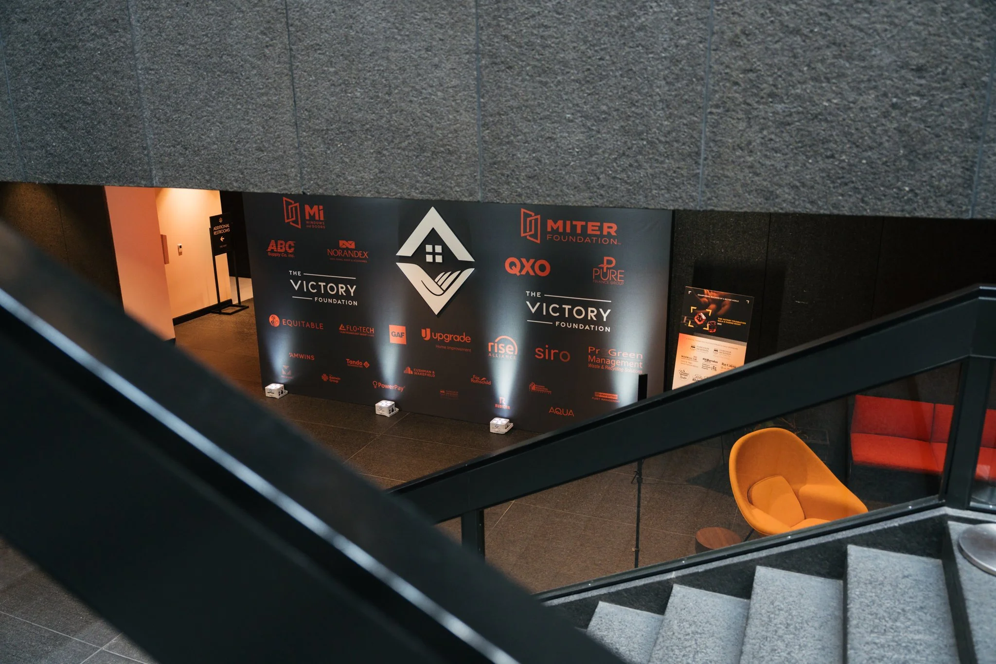 View of a staircase in a modern building, looking down into a lobby area with a large black wall displaying various company logos and the word 'Victory Foundation'. There is a single orange chair and a red chair visible on the lower level.