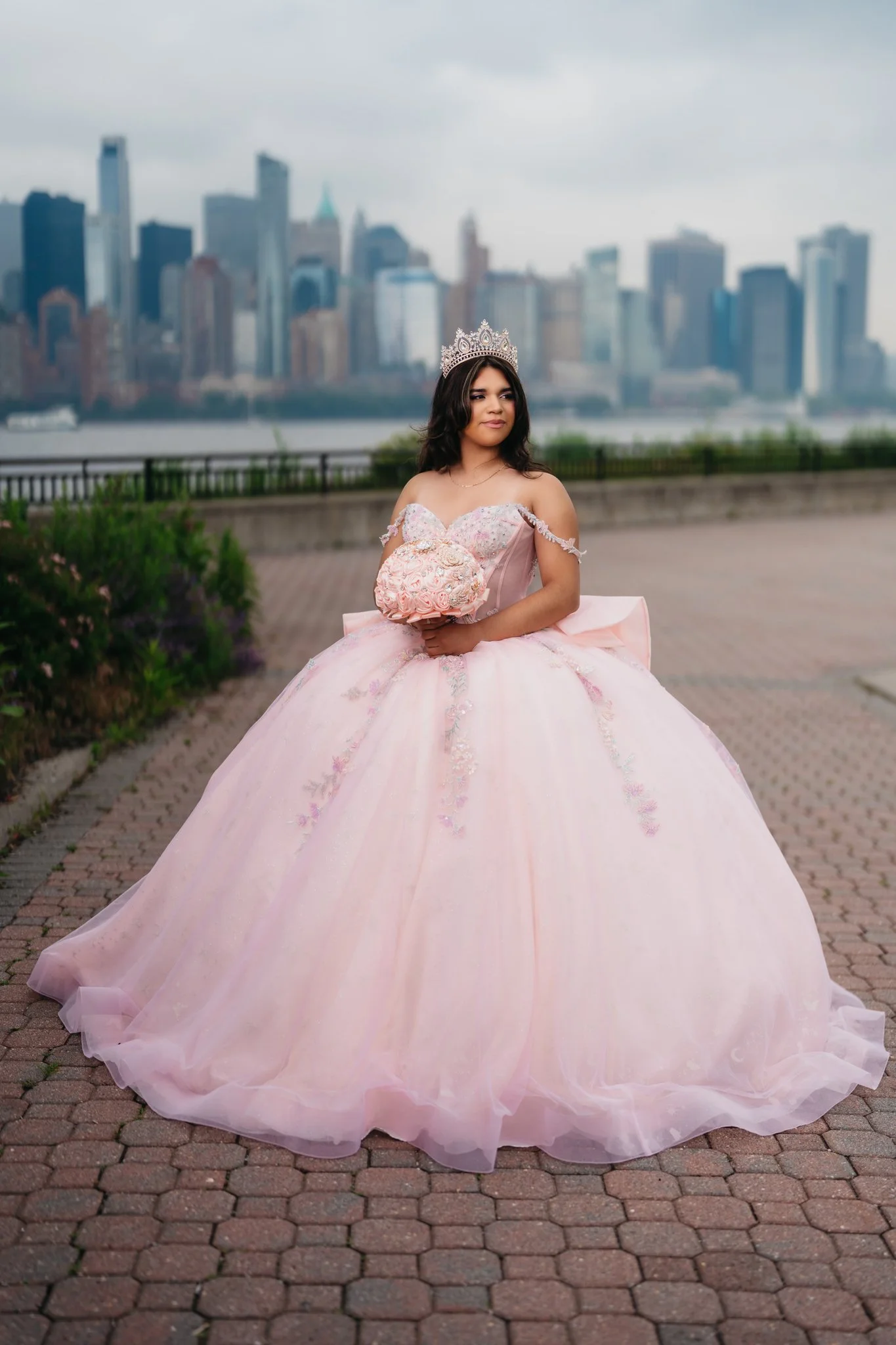 A young woman in a pink ball gown and tiara holding a bouquet of pink roses, standing outdoors with a city skyline in the background.