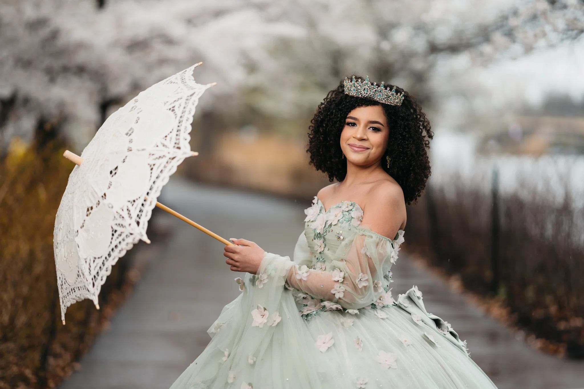 A young woman wearing a light green floral ball gown and a tiara stands outdoors holding a matching lace parasol, with a background of blurred trees and pathway.
