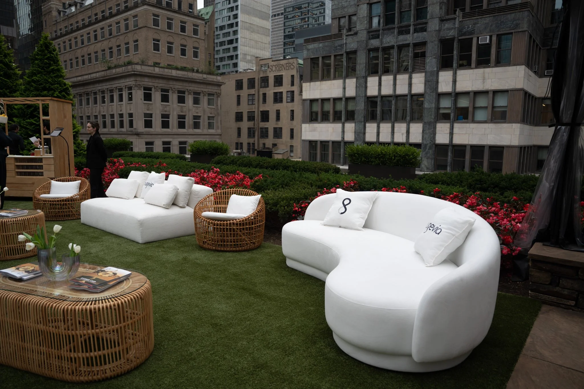 Outdoor seating area on a rooftop with white sofas, wicker chairs, and small round wicker tables on artificial grass, surrounded by pink flowers and green shrubs, with tall buildings in the background.