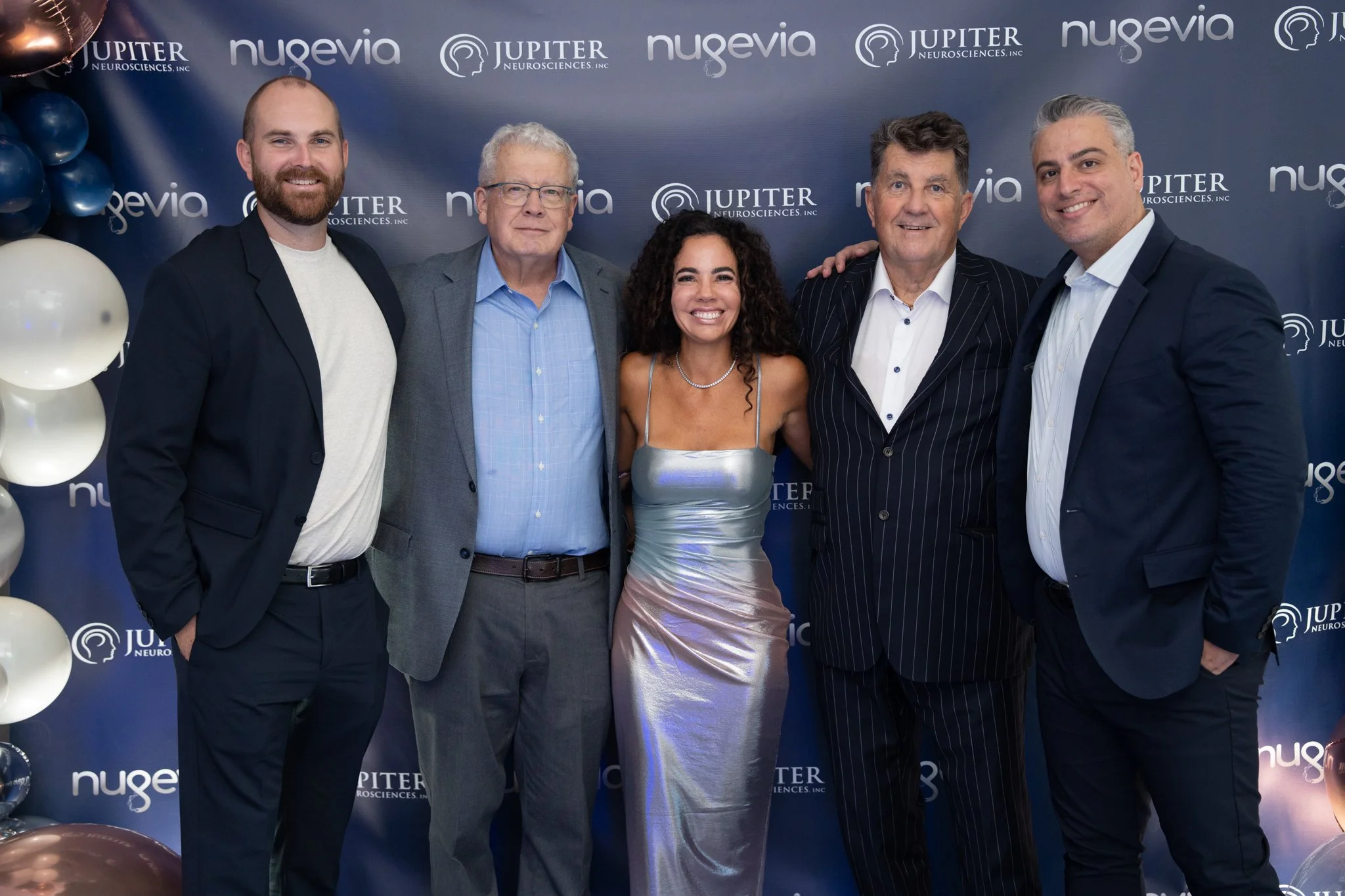 Group of five people at Nugevia and Jupiter Neurosciences event, dressed in formal attire, standing in front of a branded backdrop, smiling for the photo.