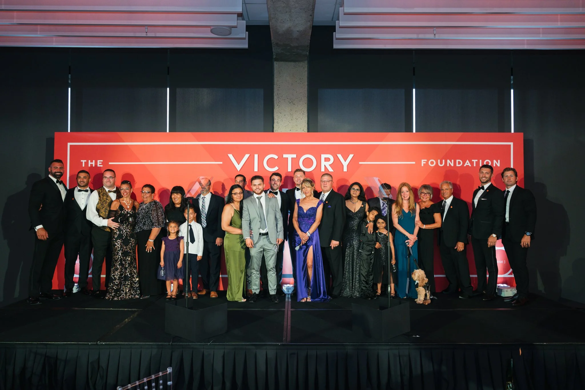 Group of people dressed in formal attire standing on stage with a red backdrop that reads 'The Victory Foundation'.