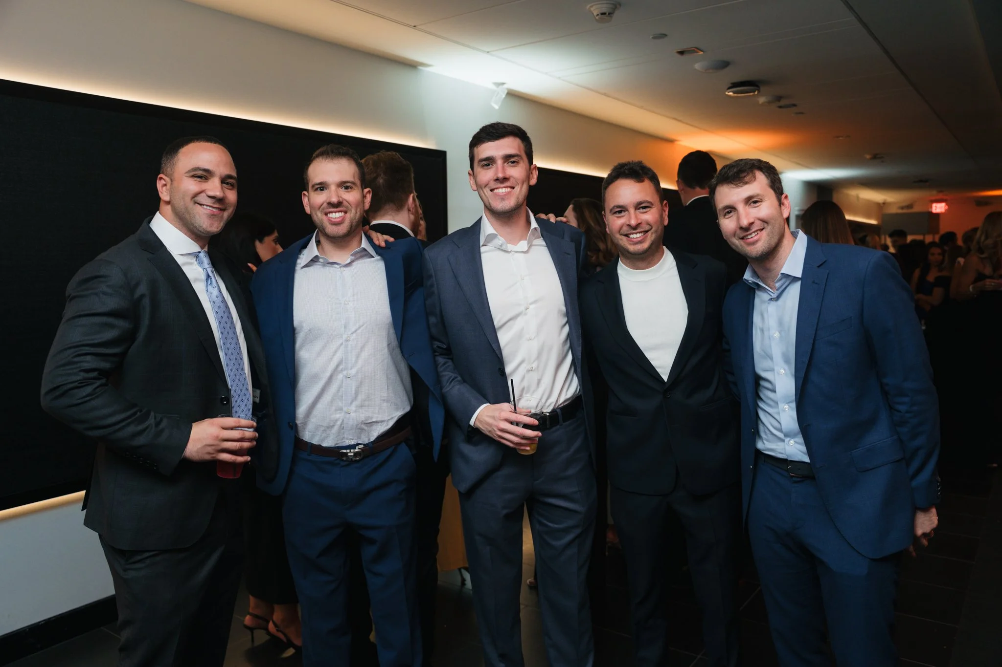 Six men in business suits standing together at an indoor social event. They are smiling and holding drinks, with a dark background and warm lighting.